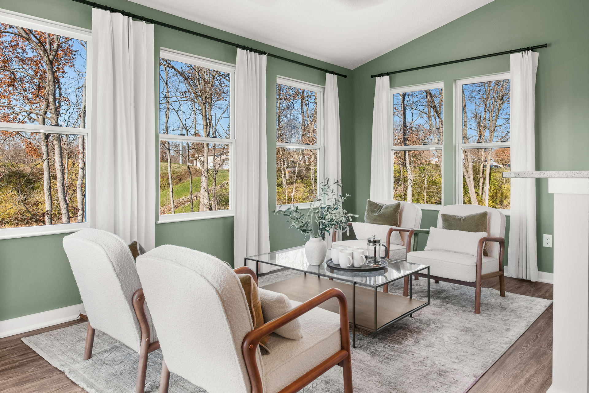 Bright and cozy living room with green walls, large windows, white curtains, and modern furniture showcasing a serene wooded view.