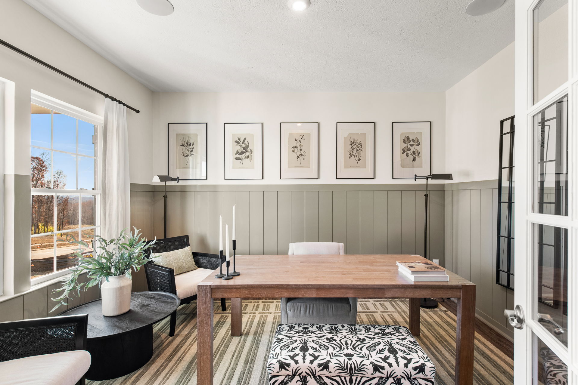 Stylish home office with botanical wall art, a wooden desk, and a cozy seating arrangement featuring a patterned ottoman and potted plant.