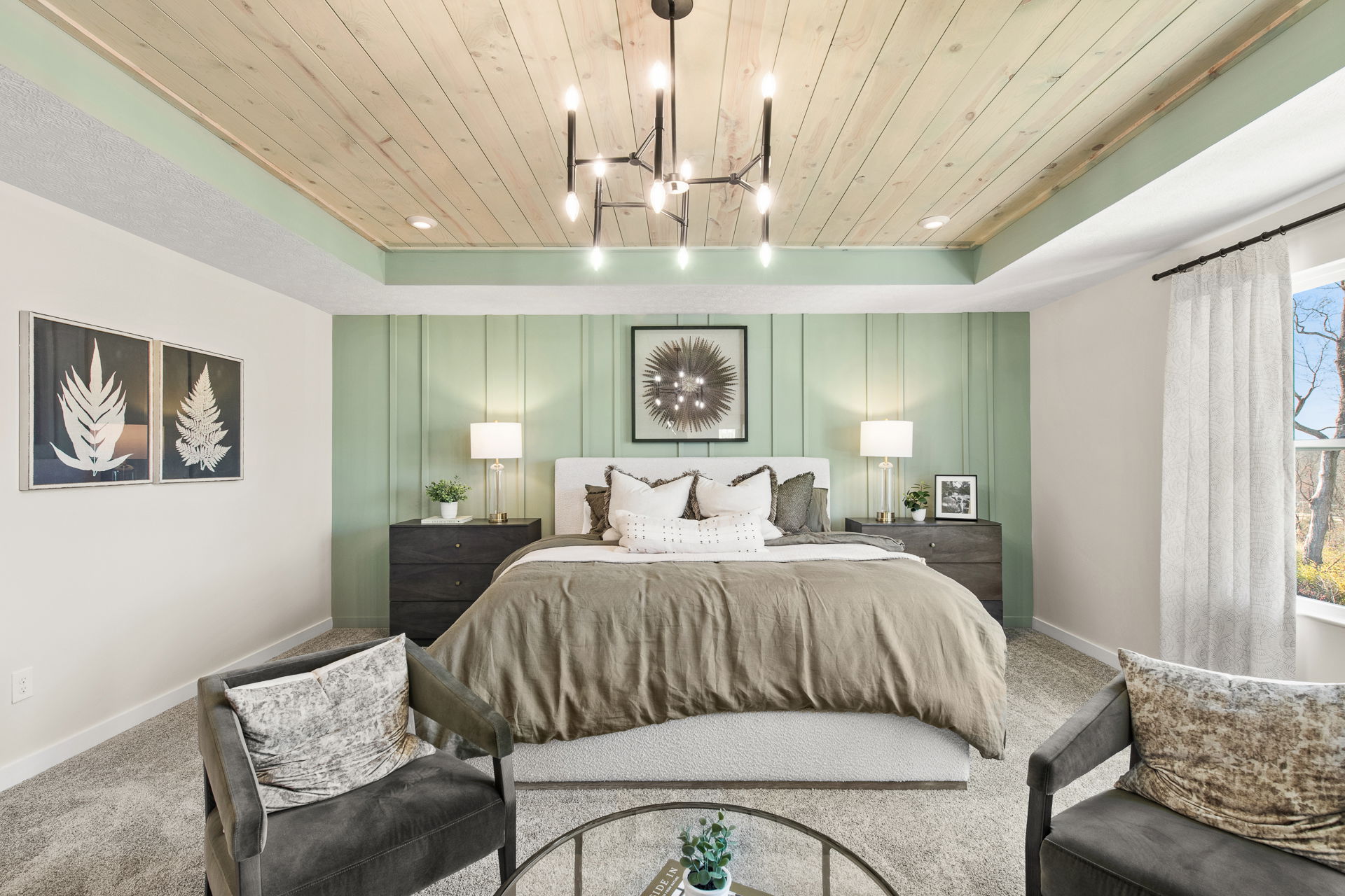 Modern bedroom with a green accent wall, cozy beige bedding, black furniture, and a stylish chandelier.