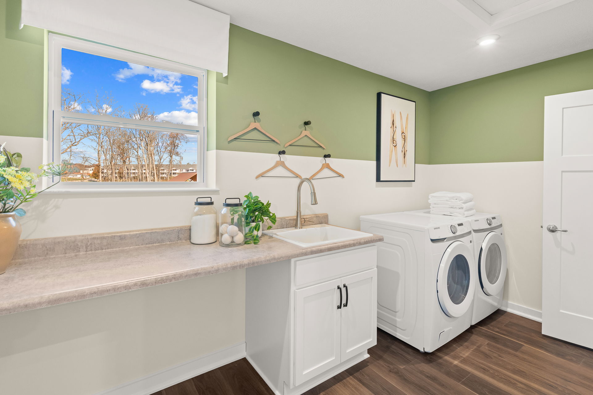 Modern laundry room with a washer, dryer, sink, countertop, and decorative elements against green and white walls, featuring a large window with a scenic view.