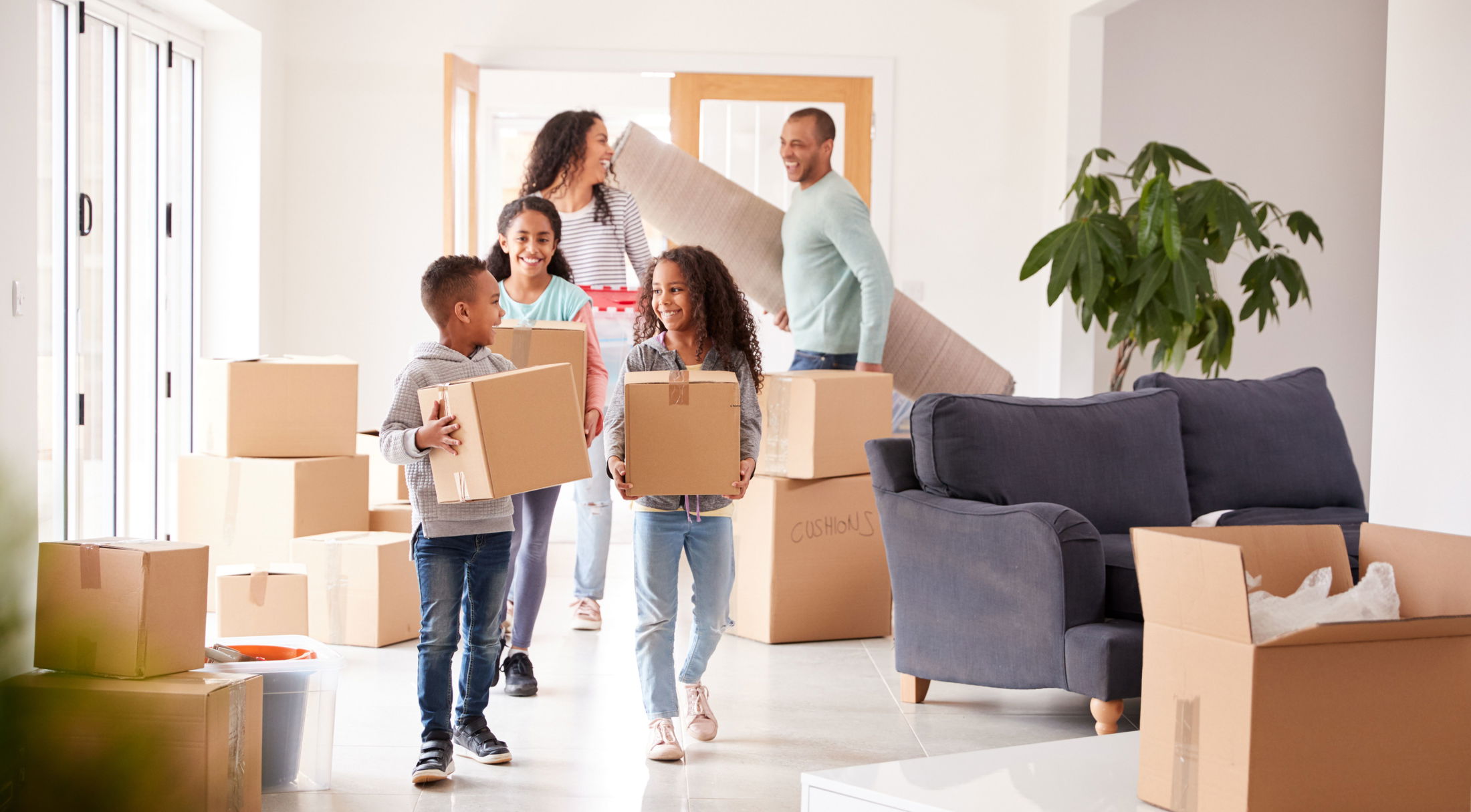 A happy family moving into a new home carrying boxes and a rug, surrounded by unpacked items.