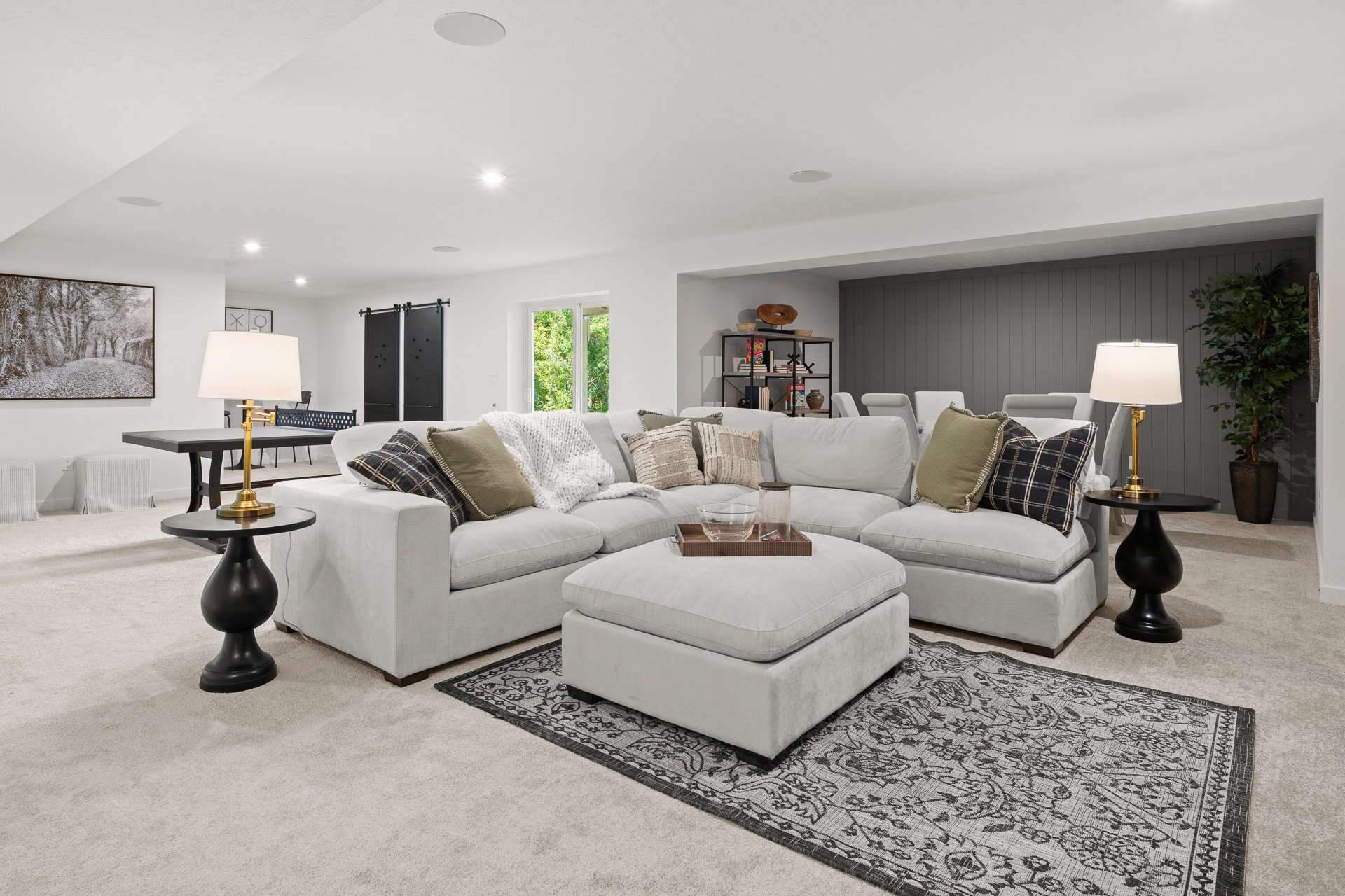 A modern living room featuring a light-colored sectional sofa, two side tables with lamps, a decorative rug, and a bookshelf in a spacious, well-lit area.