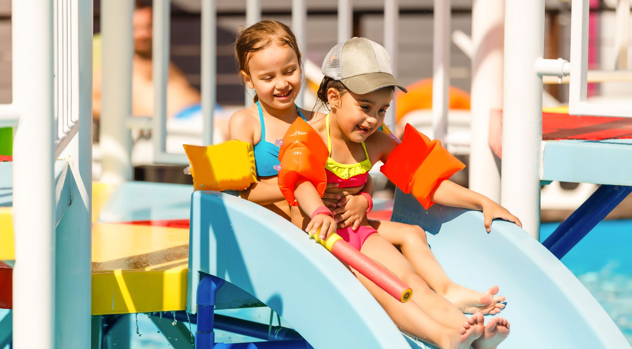 Children enjoying a water slide at the pool wearing colorful swimsuits and arm floaties.