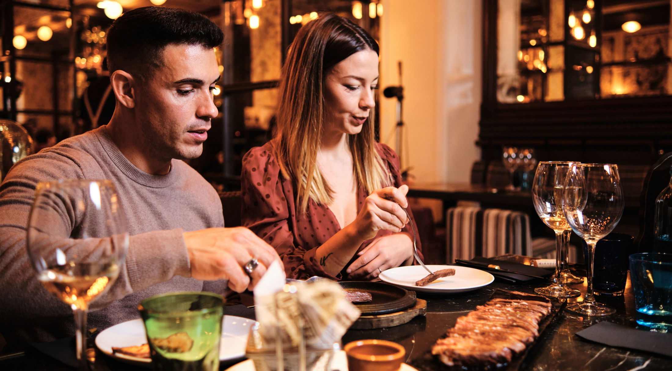 A couple enjoying a dinner of grilled meat in a warmly-lit, elegant restaurant setting.