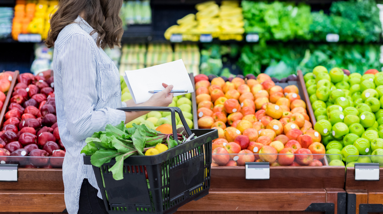 Woman shopping for fresh produce in a grocery store while holding a basket and a shopping list.