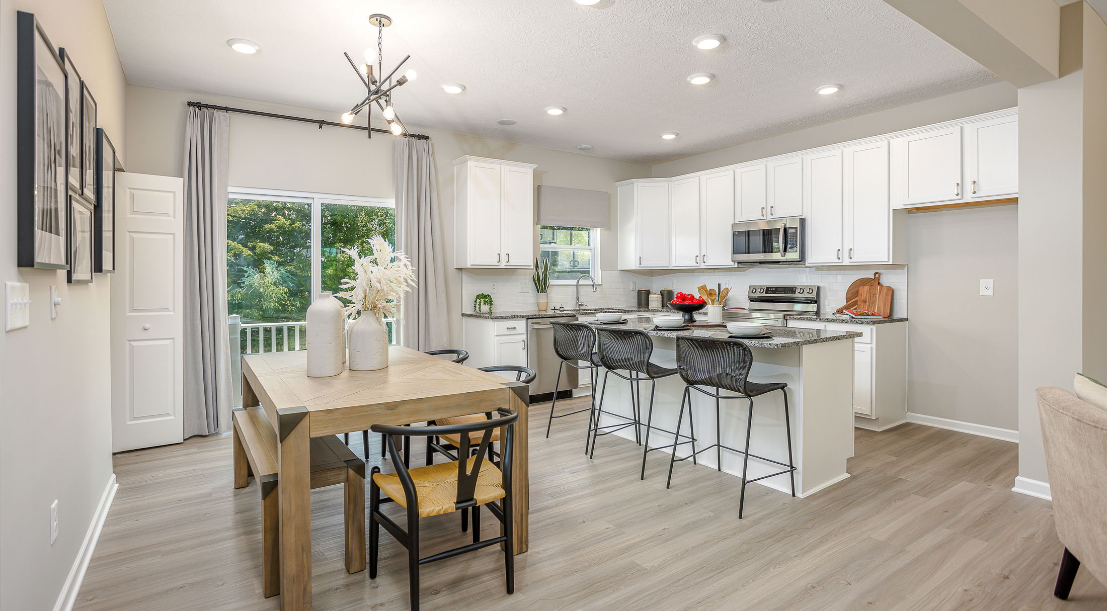 Modern kitchen and dining area with white cabinets, a wooden table, and barstools, featuring stainless steel appliances and large windows overlooking greenery.