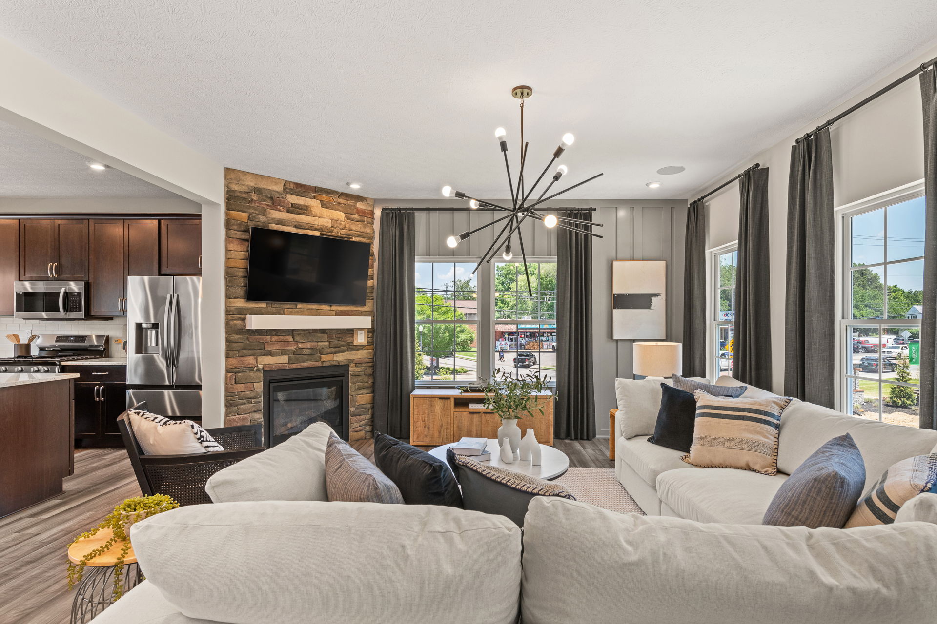 Modern living room interior with a beige sectional sofa, stone fireplace with mounted TV, stylish lighting, and open view of the kitchen.