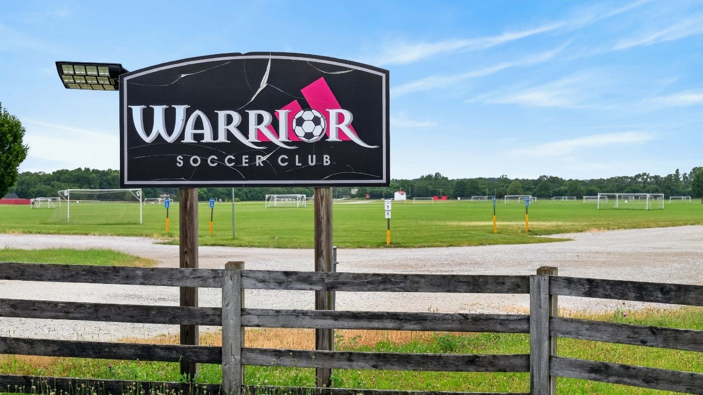 Sign for Warrior Soccer Club with a soccer ball logo and a green field and goalposts in the background.