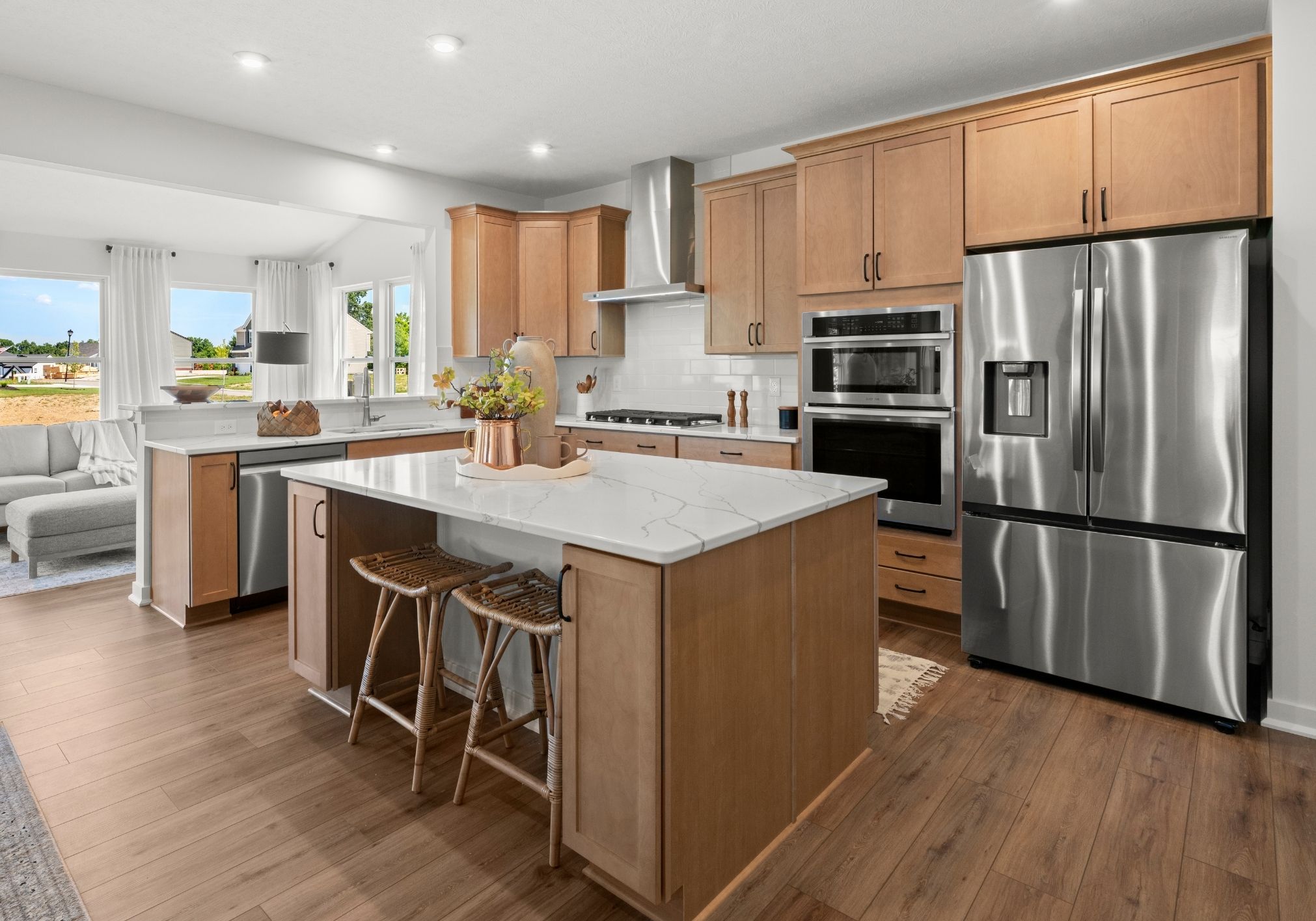 Modern kitchen featuring wooden cabinets, stainless steel appliances, a large island with bar stools, and a view of a living area.