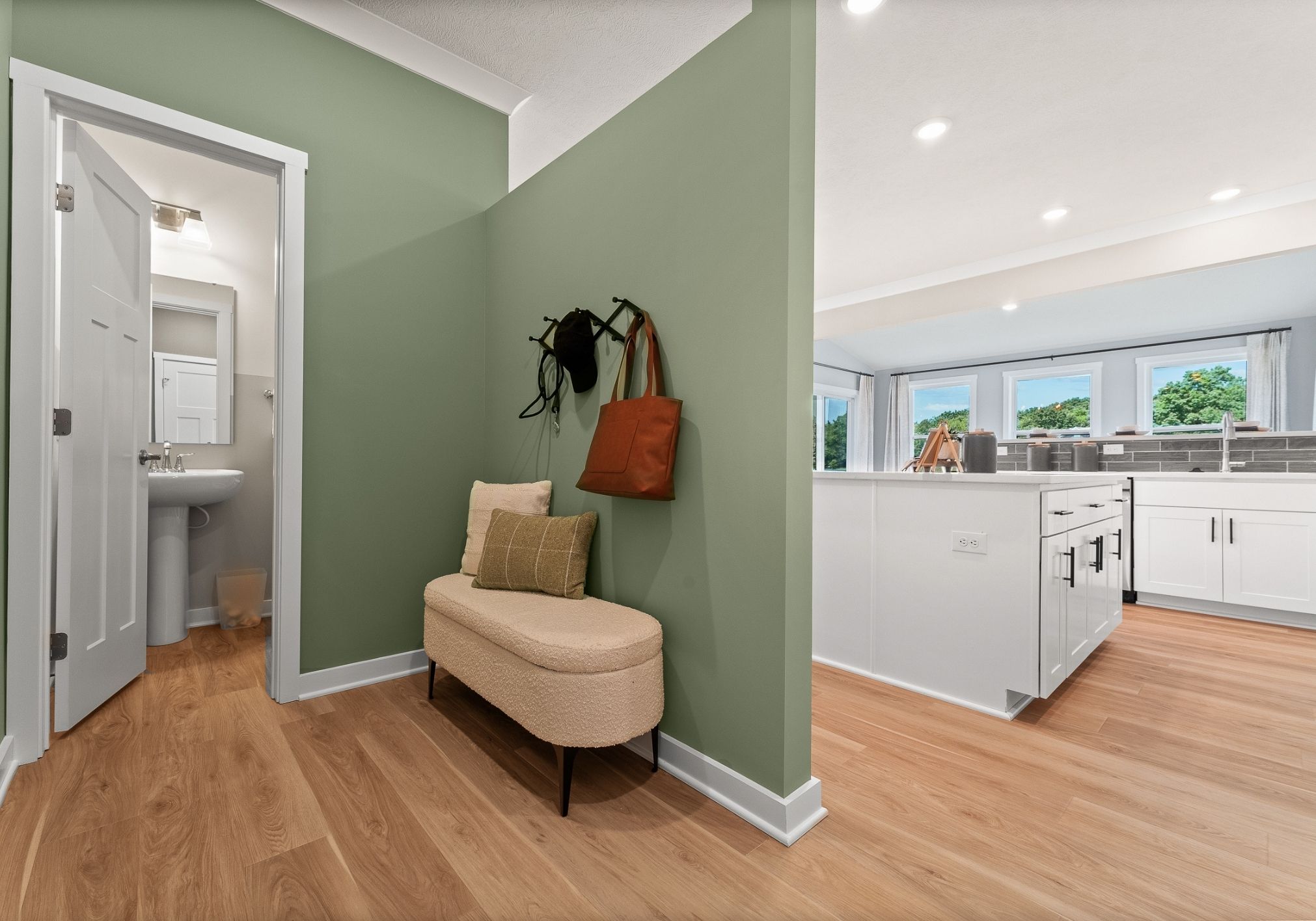 A cozy entryway with green walls featuring a small bench, a hanging bag, and a view into a brightly lit kitchen.