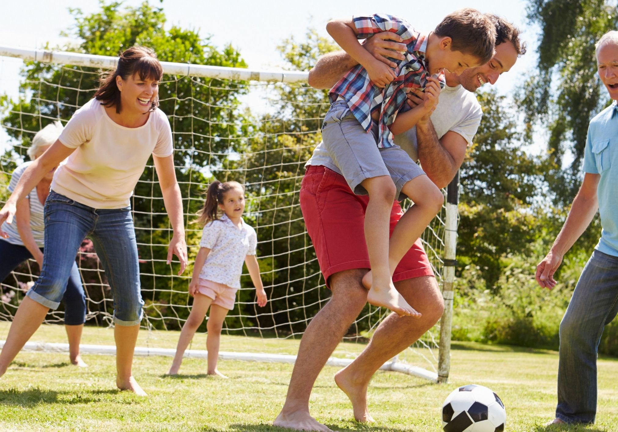A group of people joyfully playing soccer in a sunny park, with adults and children interacting around a goal and a soccer ball.