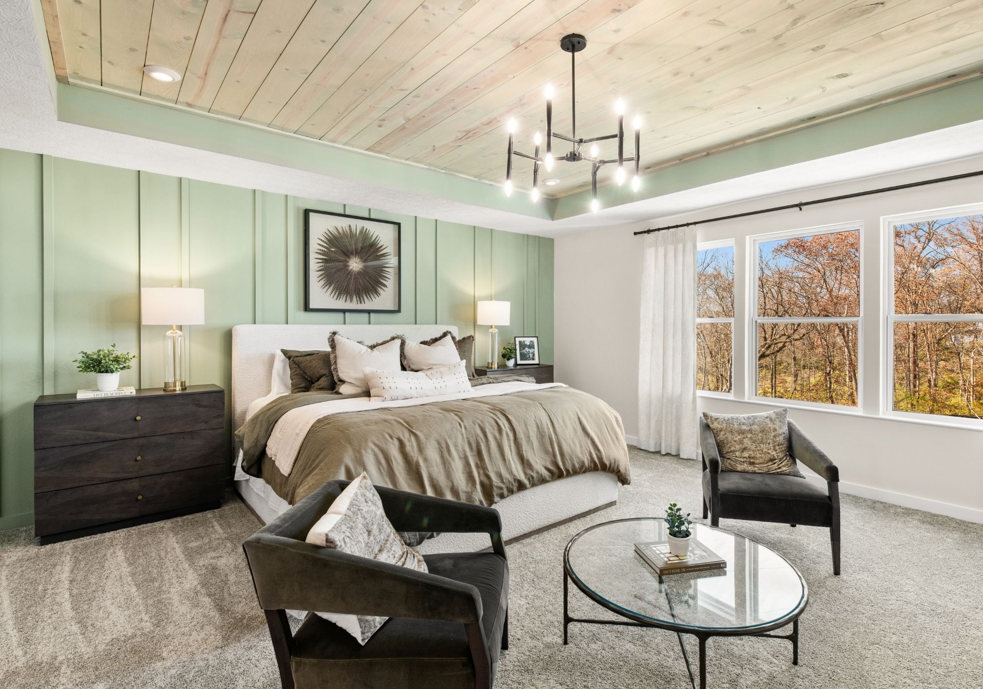 Modern bedroom featuring a green accent wall, a plush bed with neutral bedding, two black chairs, a glass coffee table, lamps, and large windows revealing a natural landscape.