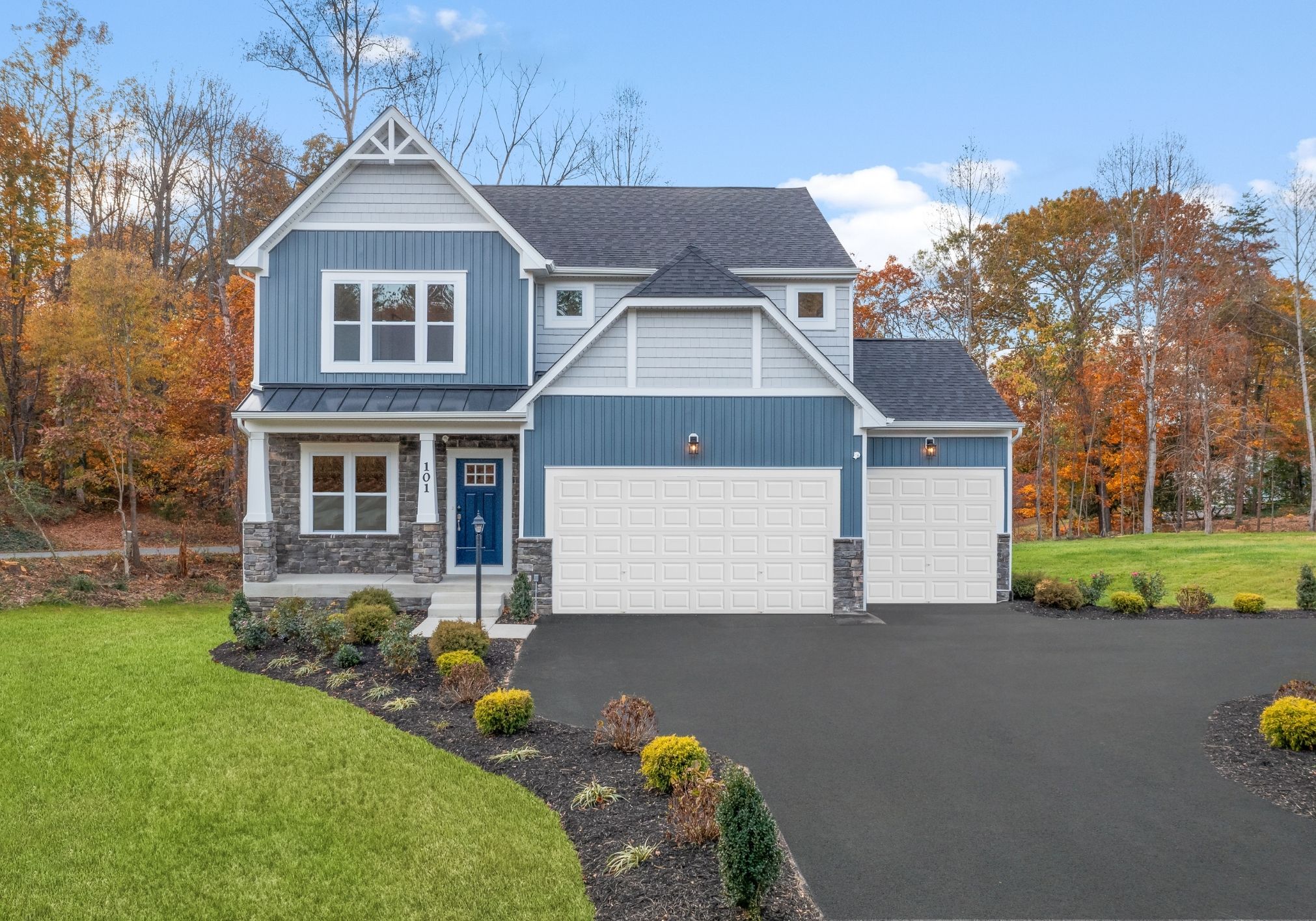 A modern blue and white house with a stone accent exterior, situated in front of trees displaying autumn foliage.