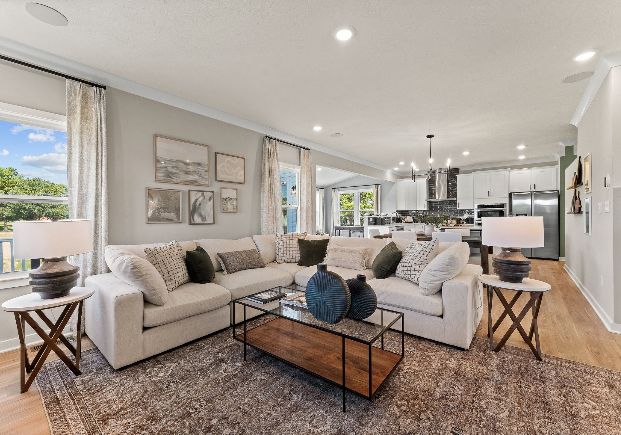 A modern living room with a white sectional sofa, decorative pillows, a coffee table, and pendant lighting, featuring a view of a kitchen with a breakfast bar.