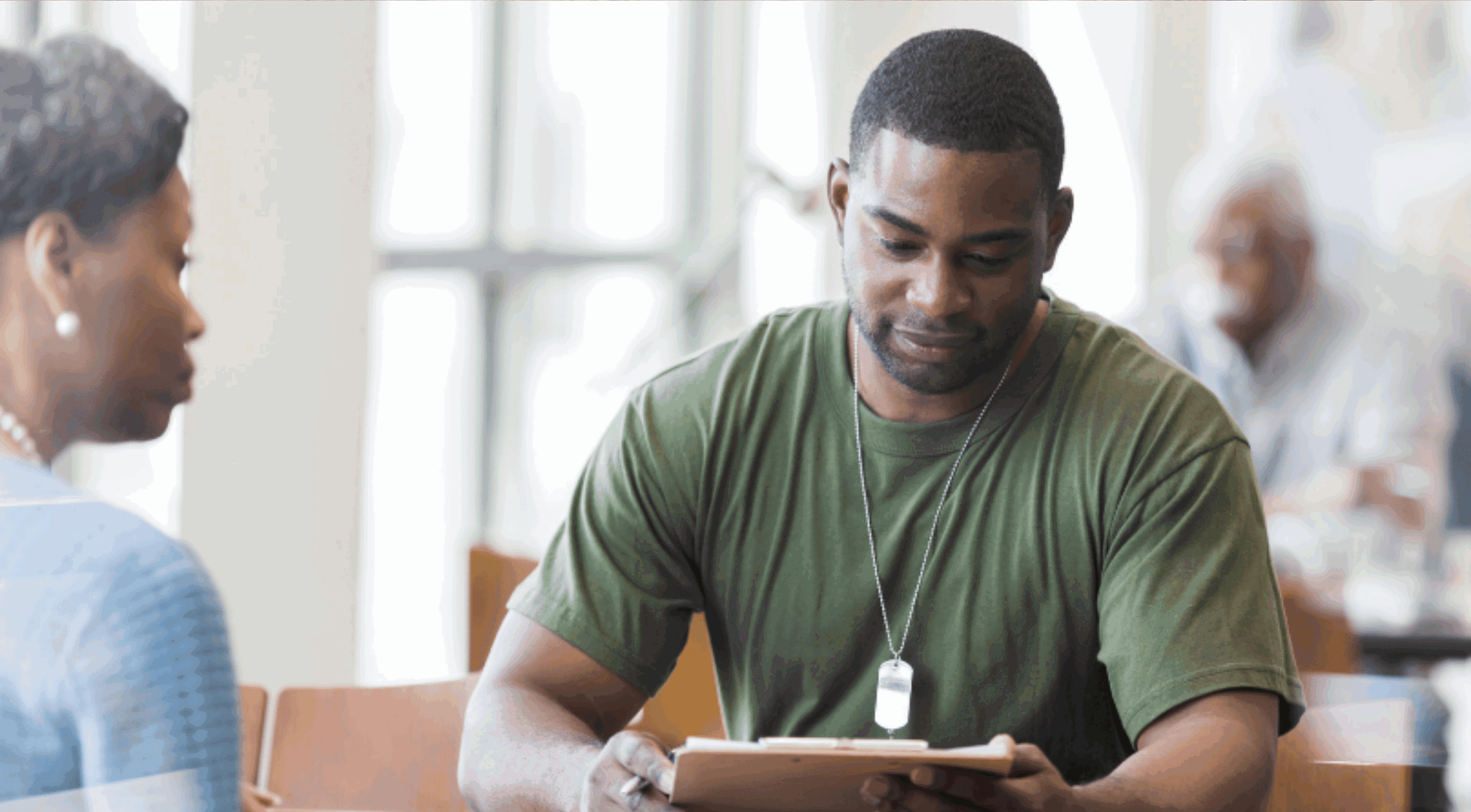 A man in a green shirt reads a clipboard during a conversation, focused inside a brightly lit room.