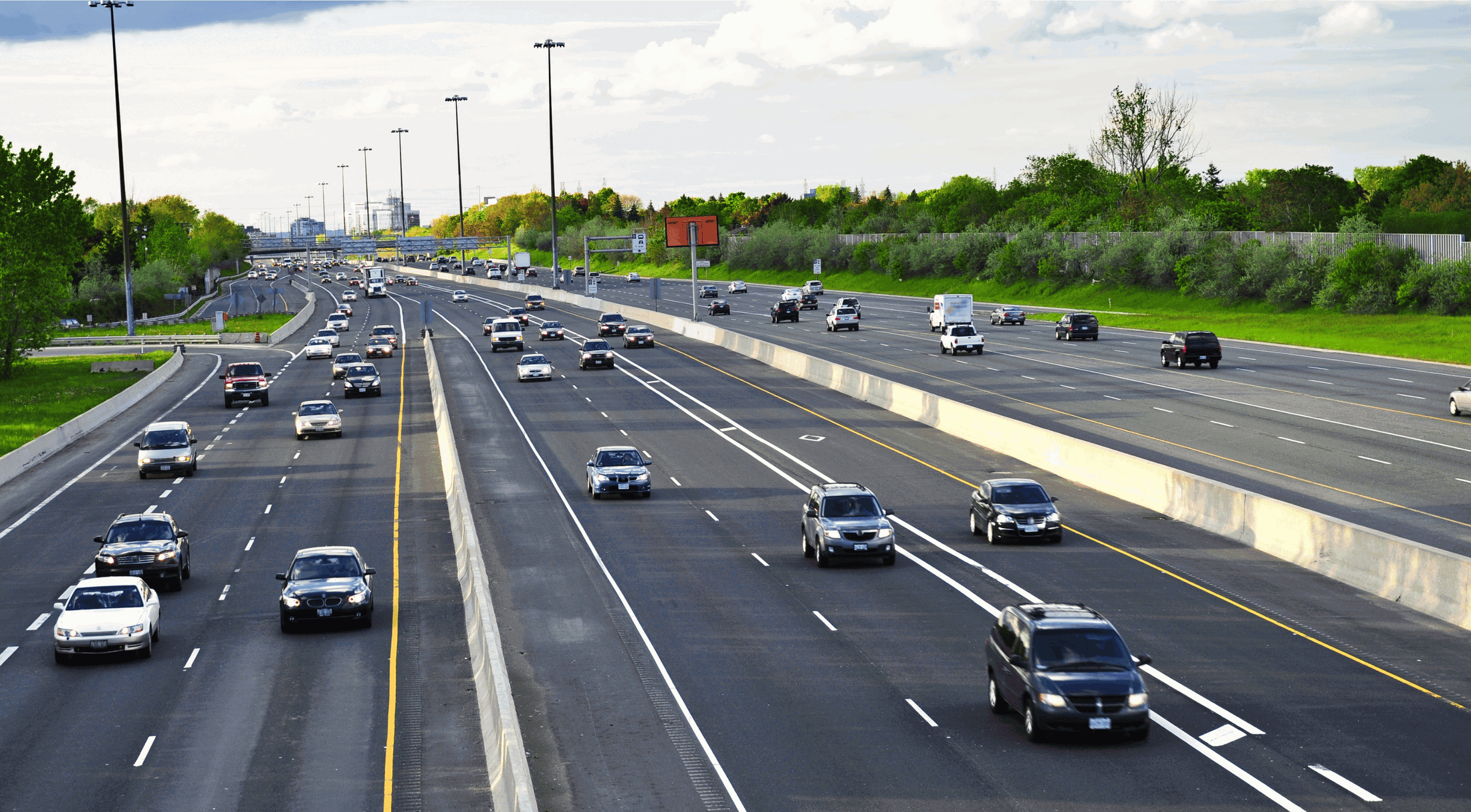 A busy multi-lane highway with cars and trucks driving in both directions, surrounded by green trees and under a partly cloudy sky.