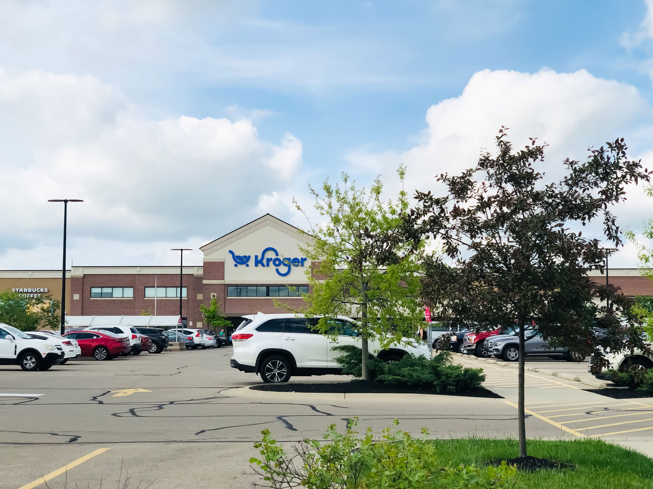 Front view of a Kroger store with a Starbucks and parking lot, featuring several cars and trees under a partly cloudy sky.