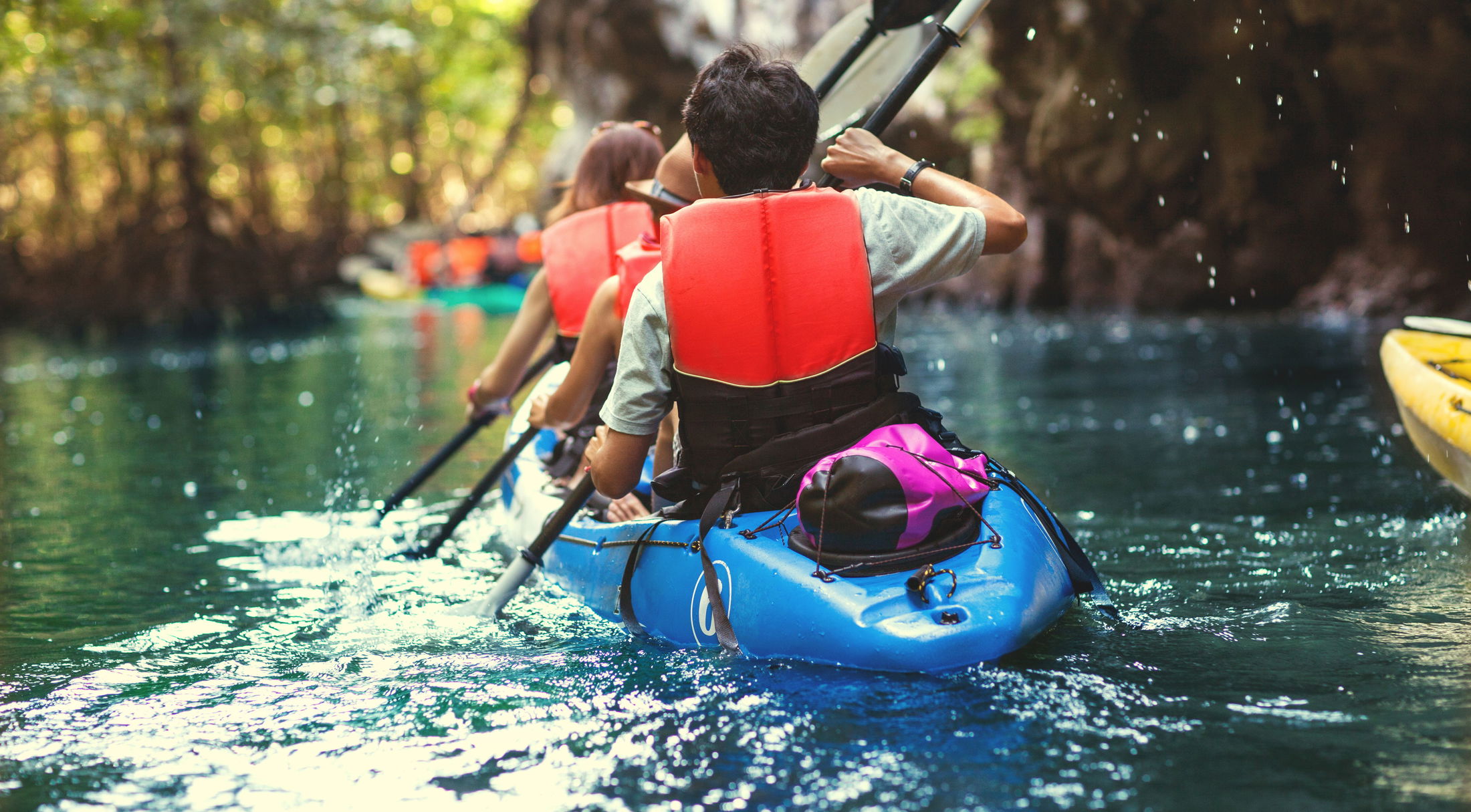 Two people kayaking through a scenic waterway surrounded by lush greenery, wearing red life vests.
