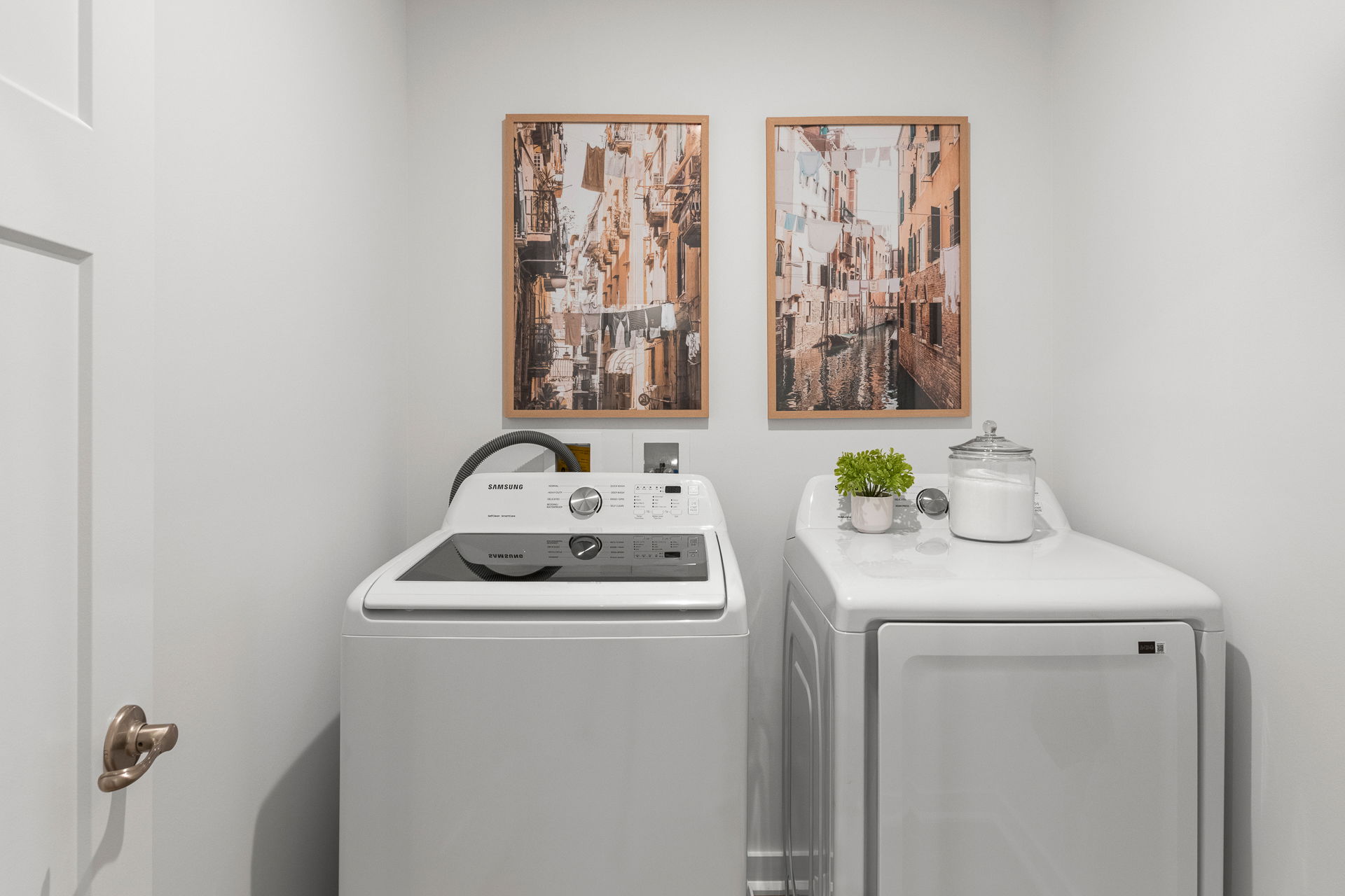 Compact laundry room with Samsung washer and dryer, decorative wall art, and a shelf holding a plant and jar.