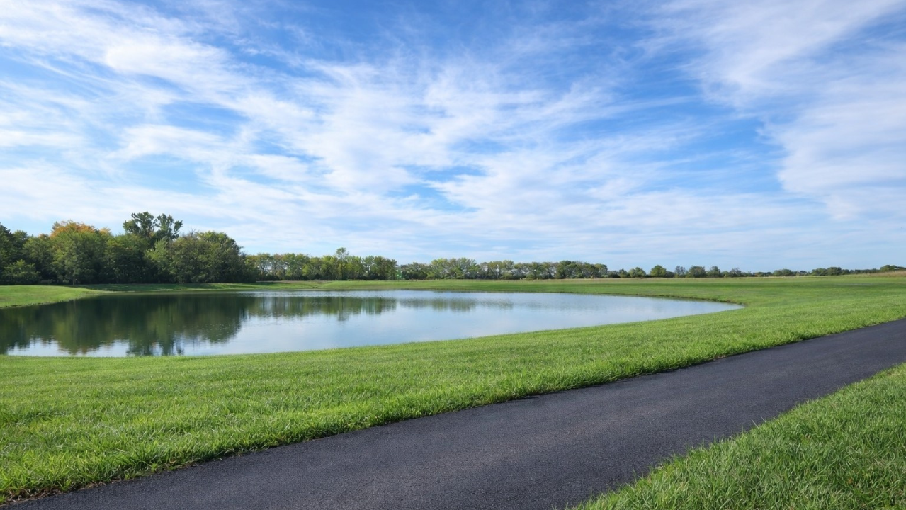 Scenic view of a calm pond surrounded by lush green grass and trees under a blue sky with white clouds.