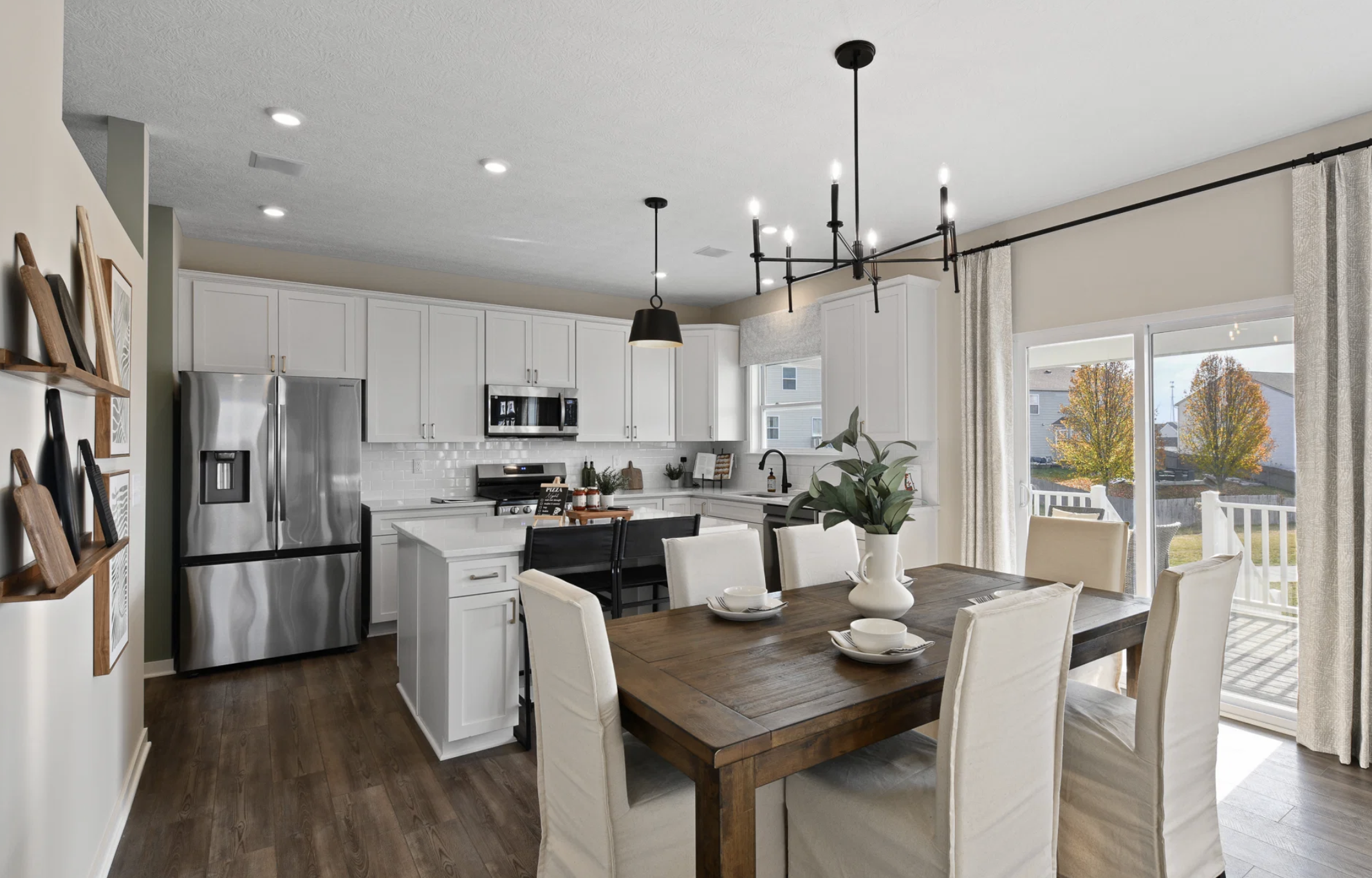 Modern kitchen and dining area featuring white cabinets, stainless steel appliances, a wooden dining table, and large windows with a view of trees outside.