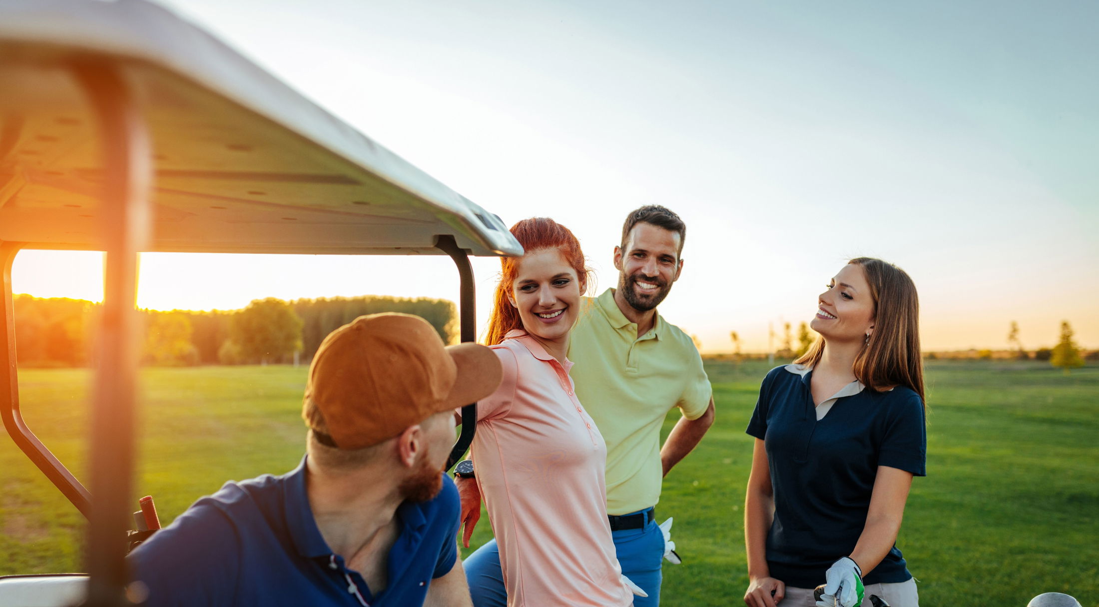 A group of four friends enjoying a day of golf and laughing near a golf cart at sunset.