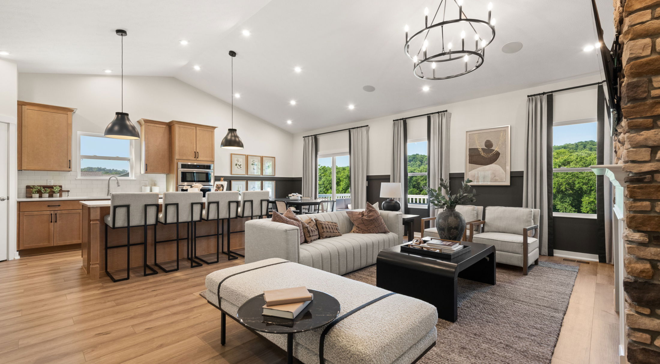 Modern open-concept living room and kitchen with neutral tones, large windows, and rustic accents like pendant lighting and stone fireplace.