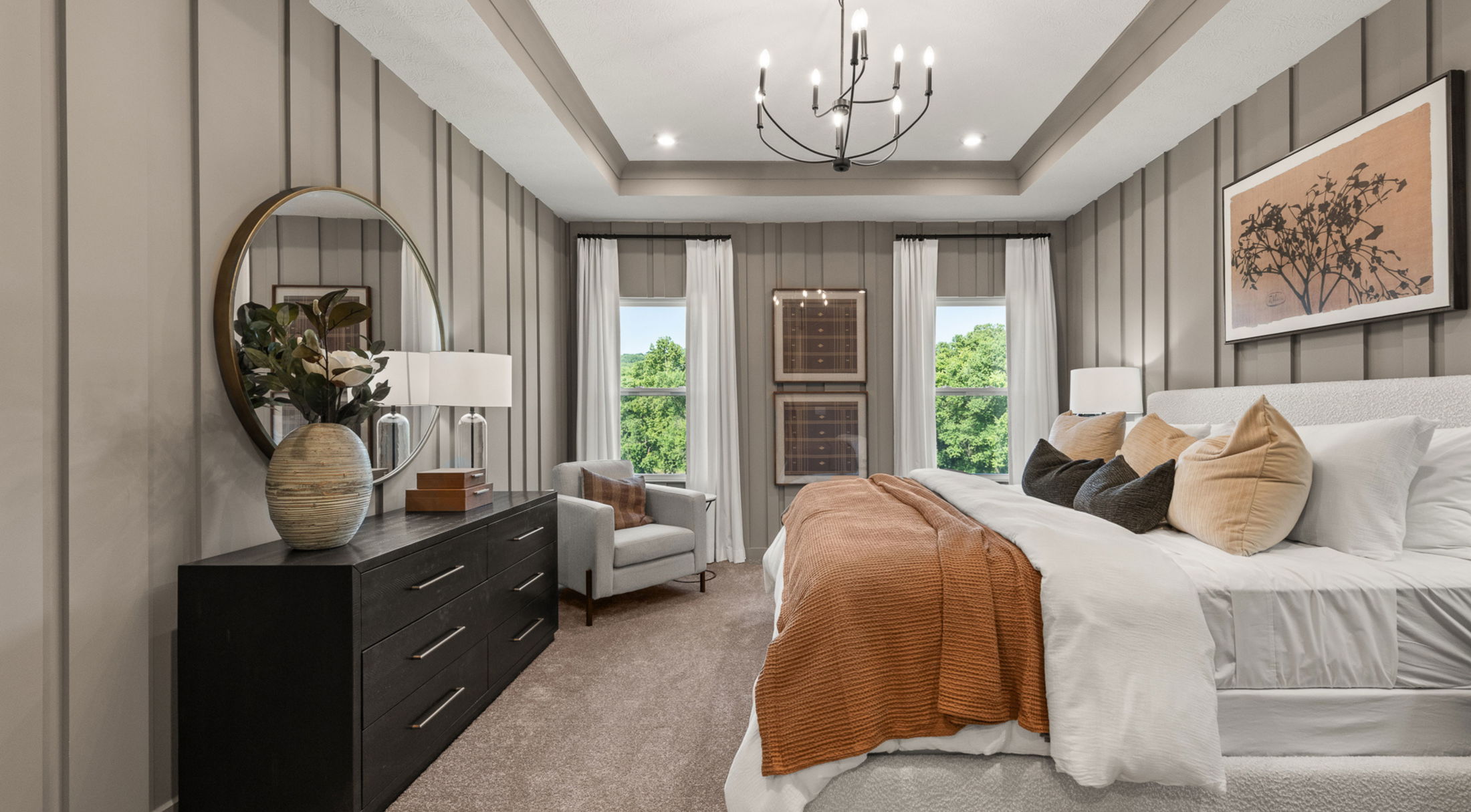 Elegant bedroom with neutral decor, featuring a cozy bed, textured cushions, large mirror above a dark dresser, modern chandelier, gray armchair, and artwork against paneled walls.