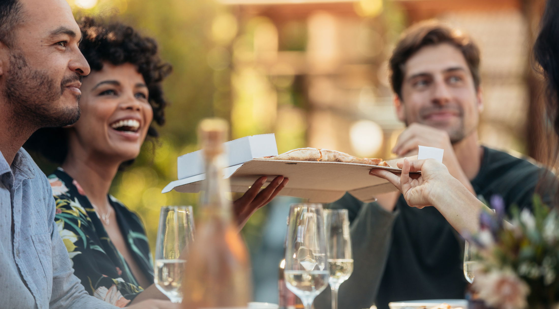 A group of friends enjoying pizza and wine outdoors, smiling and socializing in a relaxed setting.