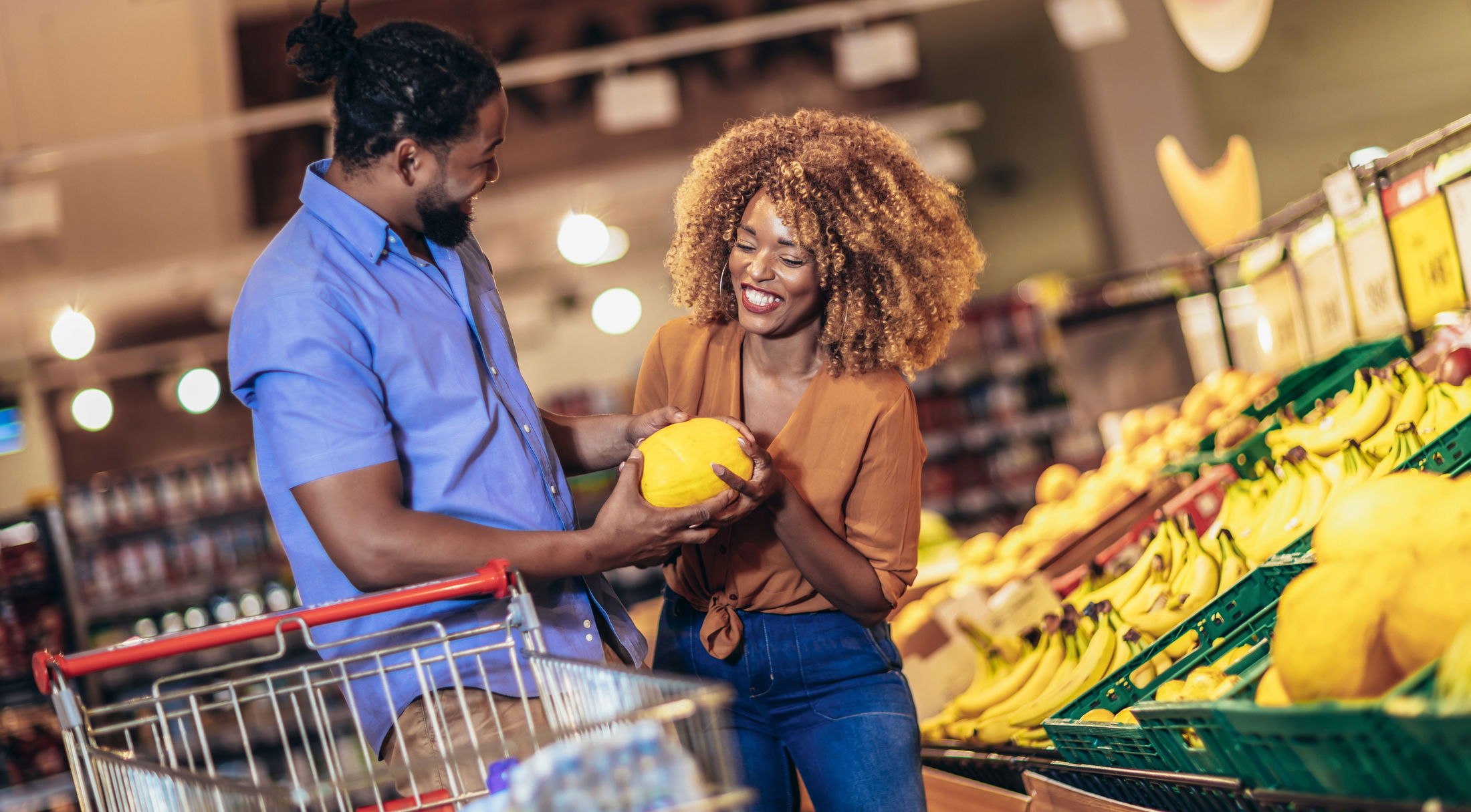 A couple shopping for fresh produce, smiling and holding a melon in a grocery store aisle filled with bananas and other fruits.