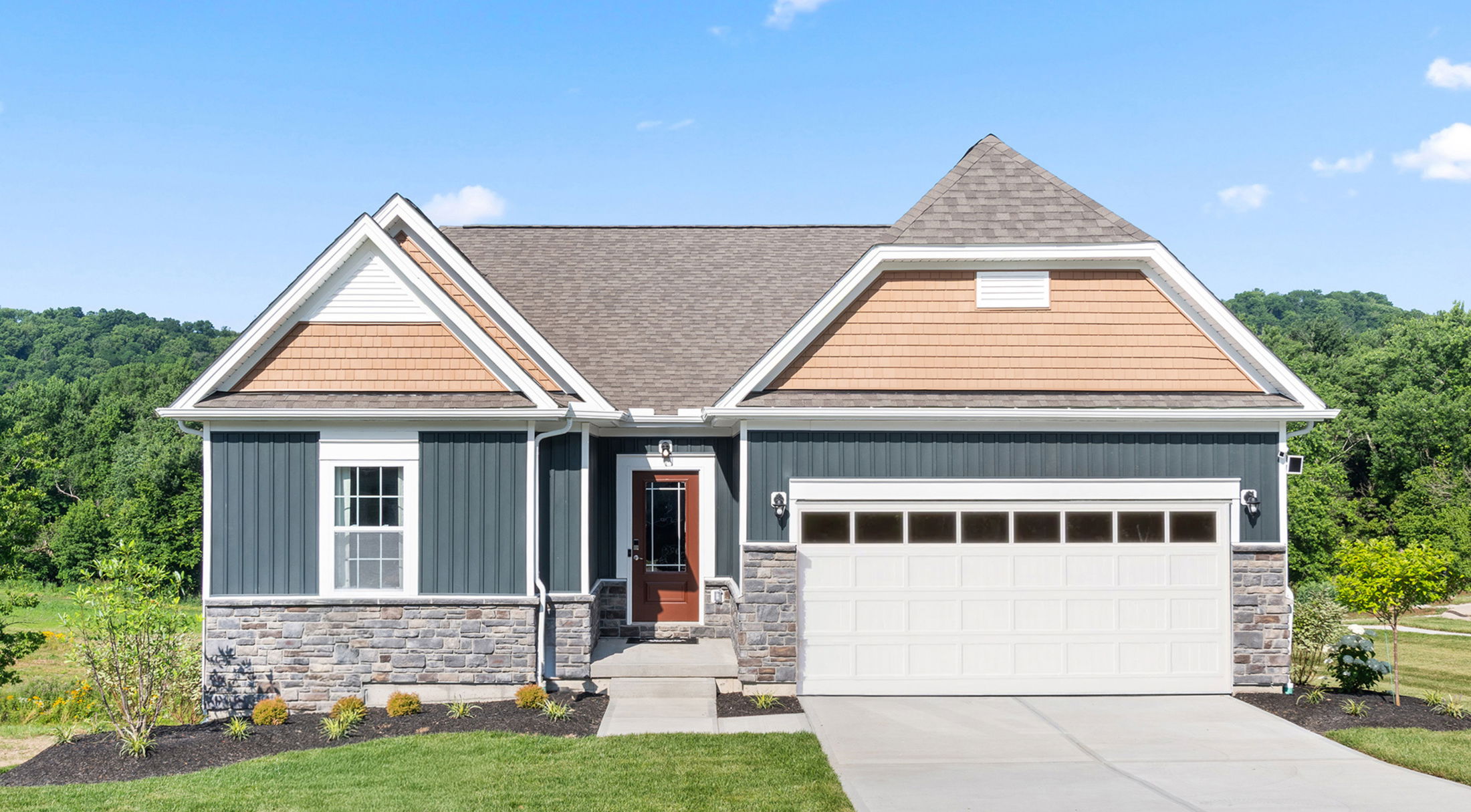 Modern single-family home with blue siding, stone accents, and a two-car garage set against a lush green landscape.