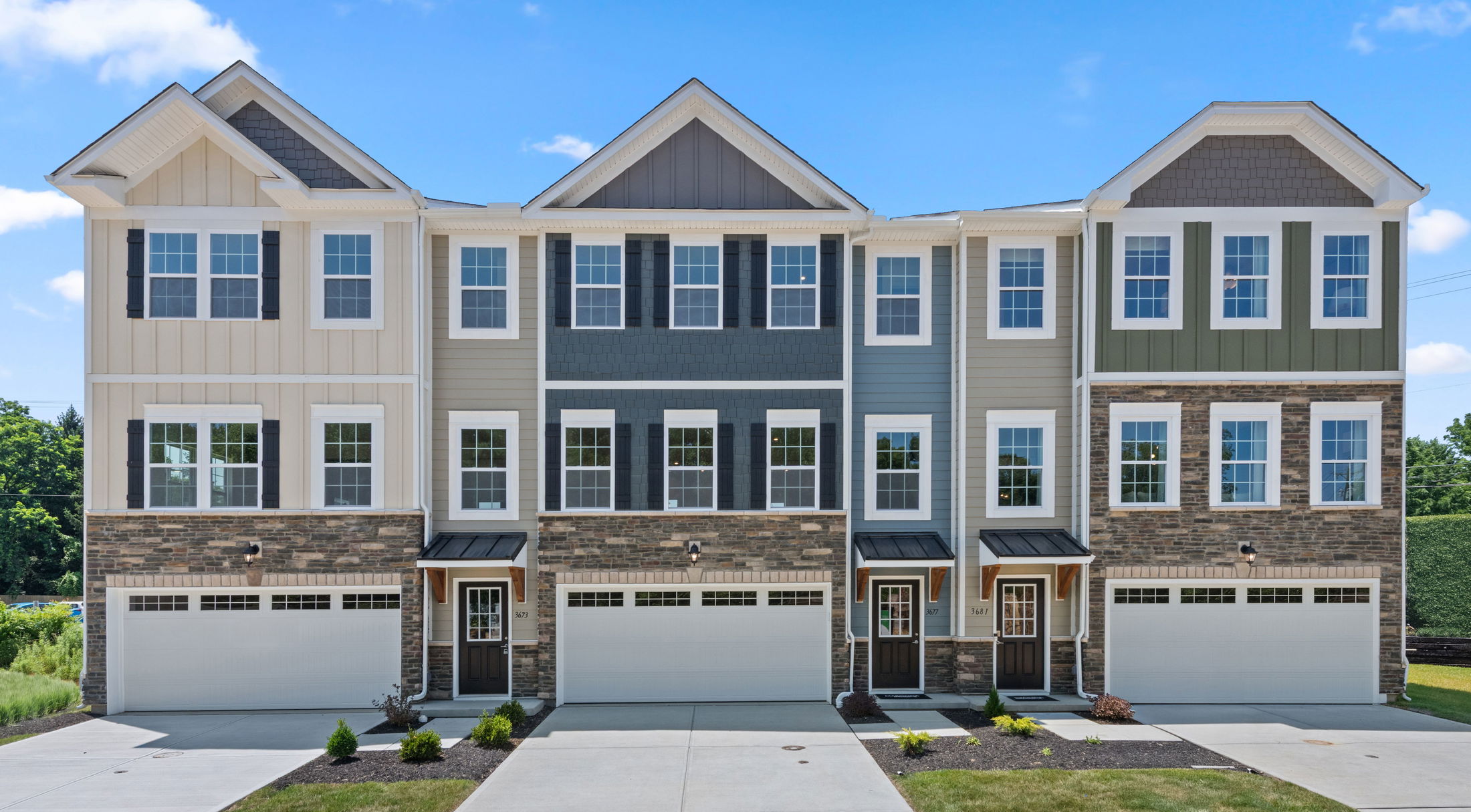 Front view of modern townhouse with stone and siding exterior, showcasing garage doors and numerous windows under a clear blue sky.