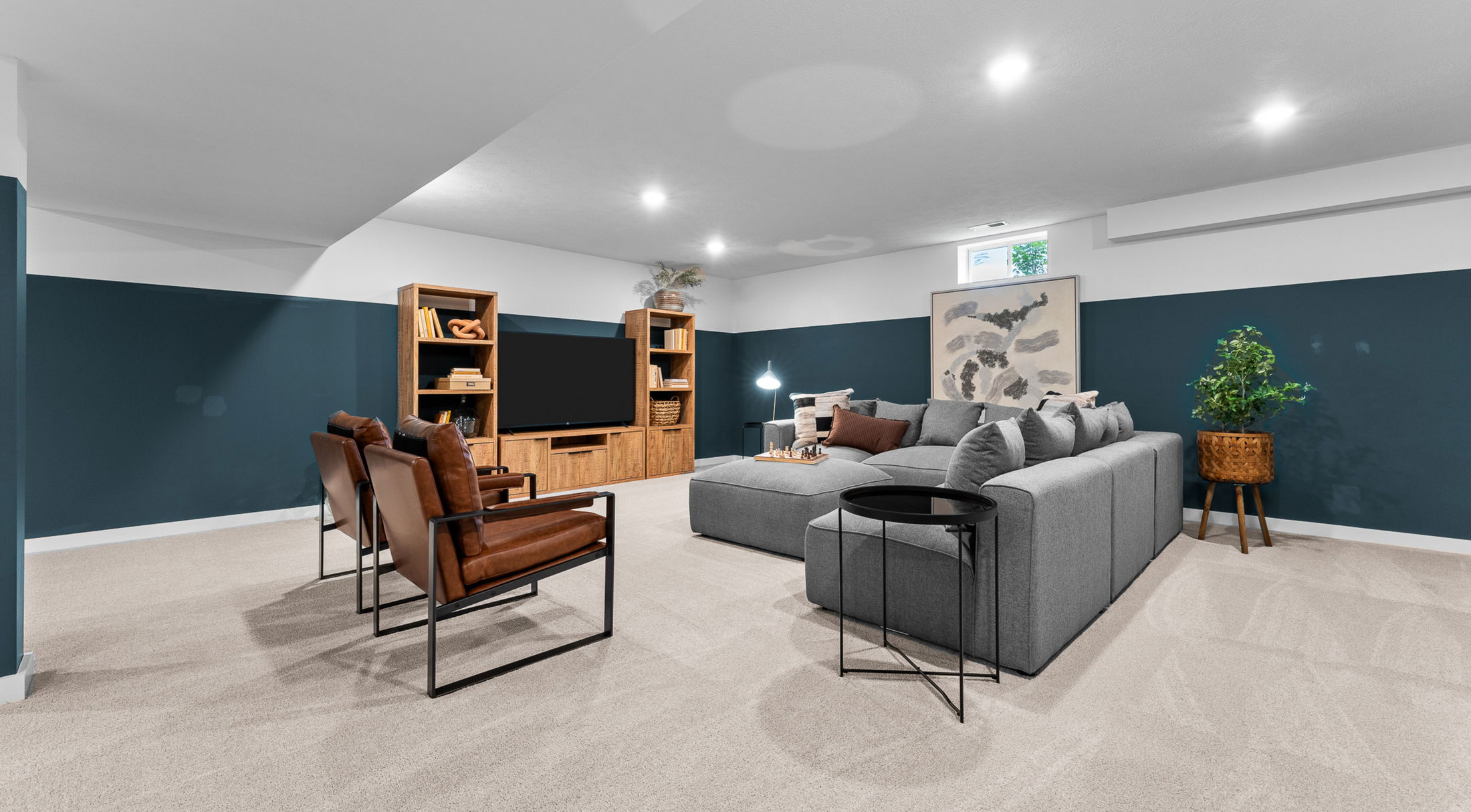 Stylish basement media room with gray sectional sofa, brown leather chairs, large TV, dark teal accent walls, and modern decor.