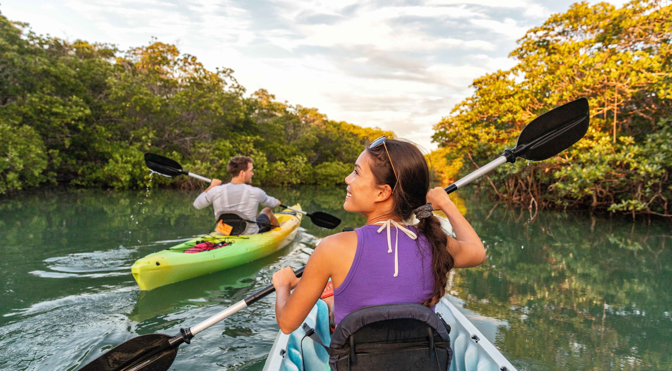 Two people kayaking through a calm, scenic mangrove forest under a clear sky.