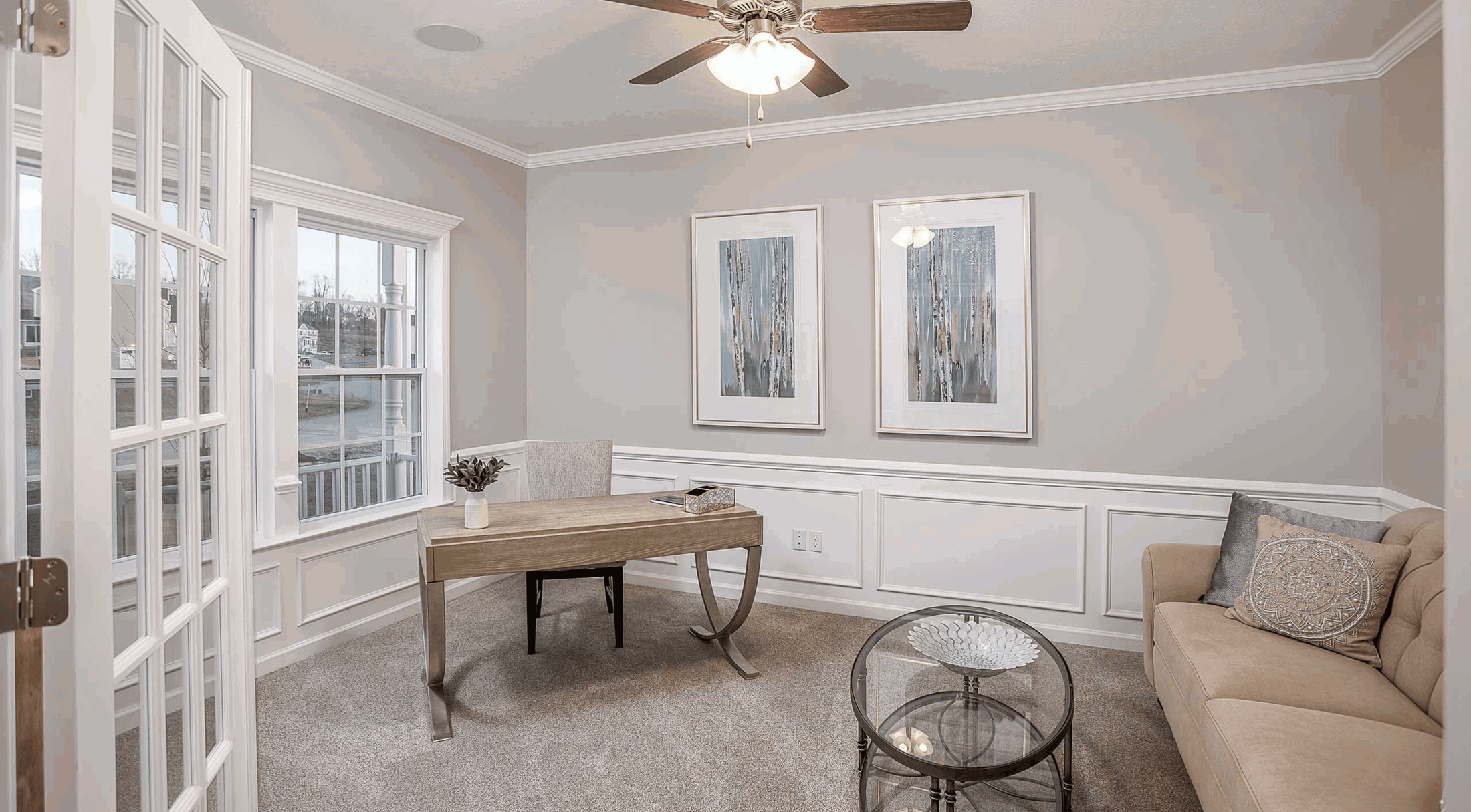 Modern home office with a wooden desk, beige sofa, glass coffee table, and elegant decor in a bright, neutral-toned room.