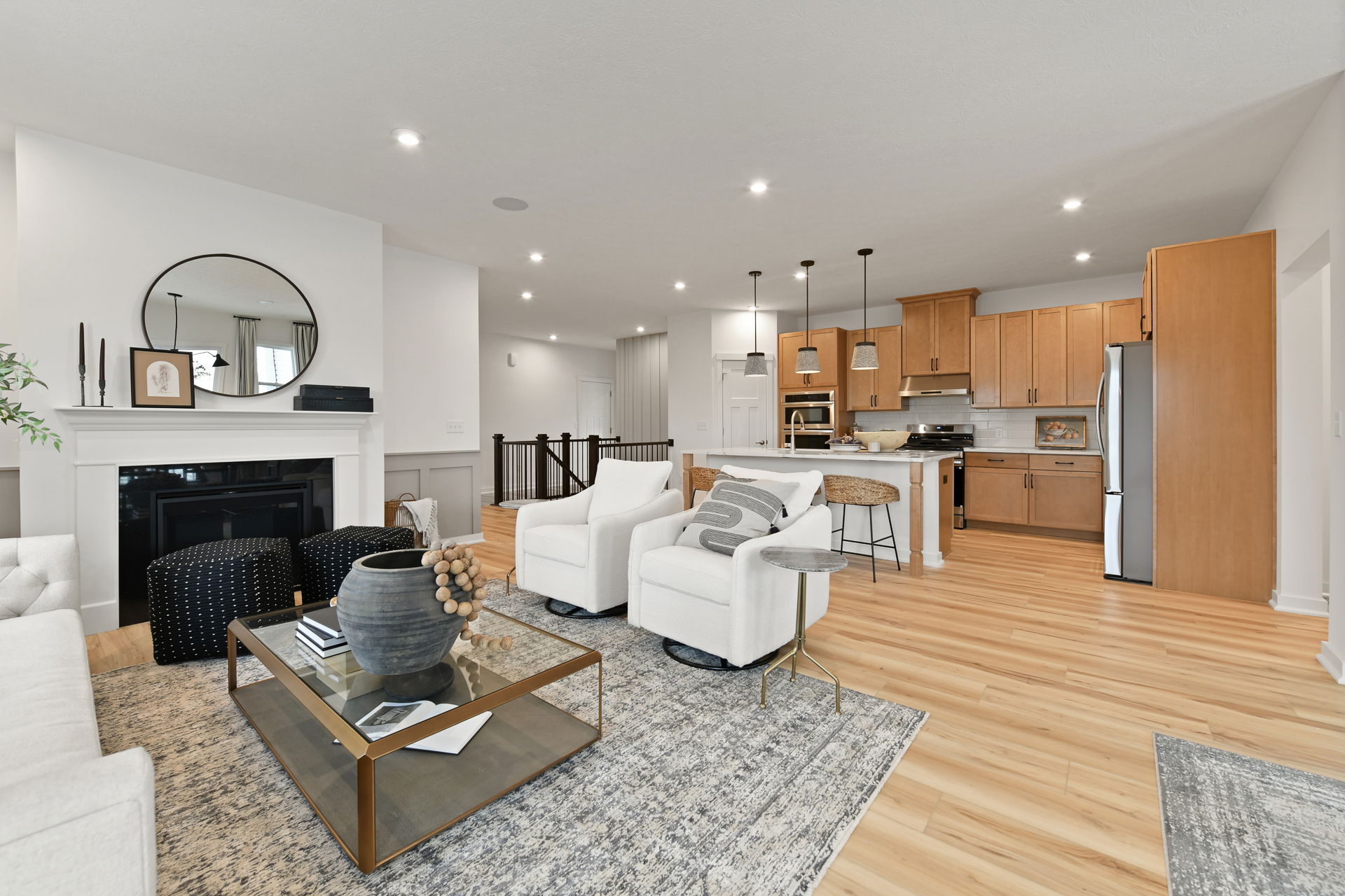 Modern open-concept living room and kitchen with light wood floors, cozy white chairs, and a decorative fireplace.