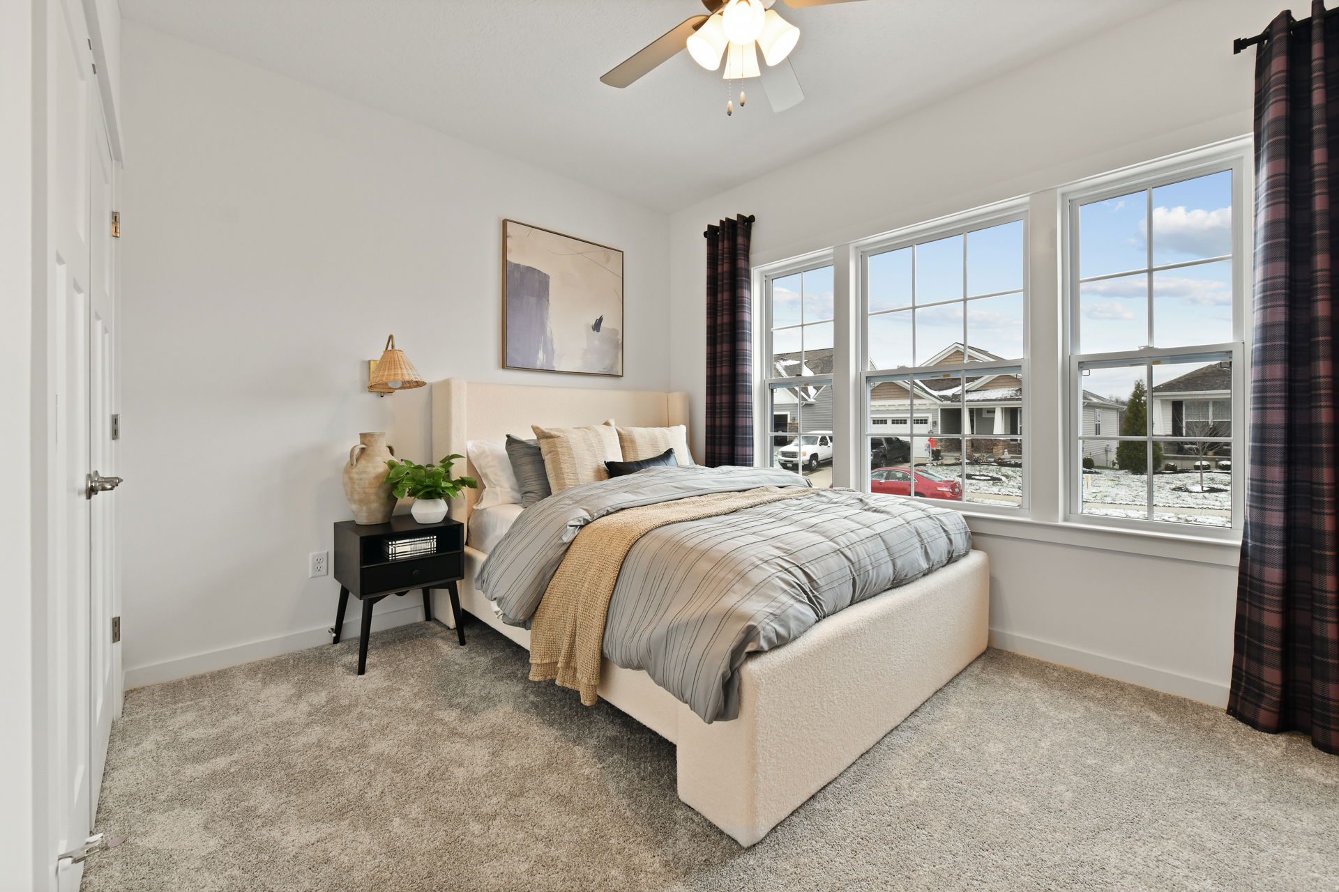 Modern bedroom with beige upholstered bed, striped bedding, and large windows overlooking a suburban neighborhood.