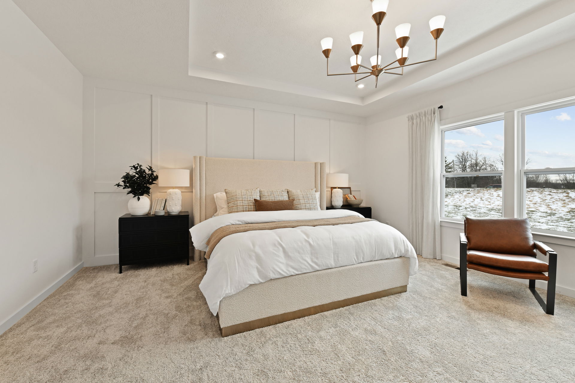 Modern bedroom interior with a beige upholstered bed, stylish lighting fixture, and a large window offering an outdoor view.