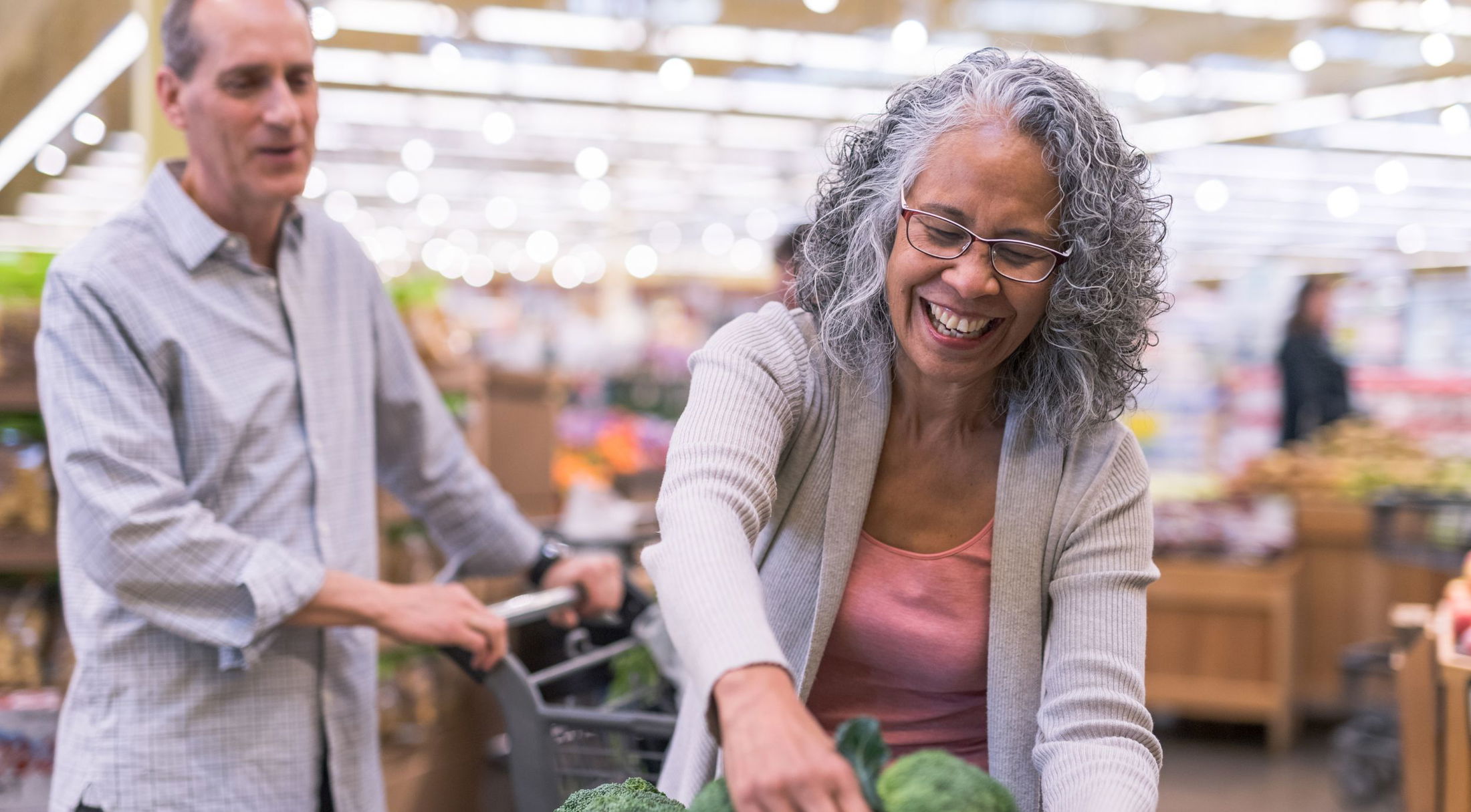 An elderly couple happily shops for fresh vegetables at a brightly lit grocery store.