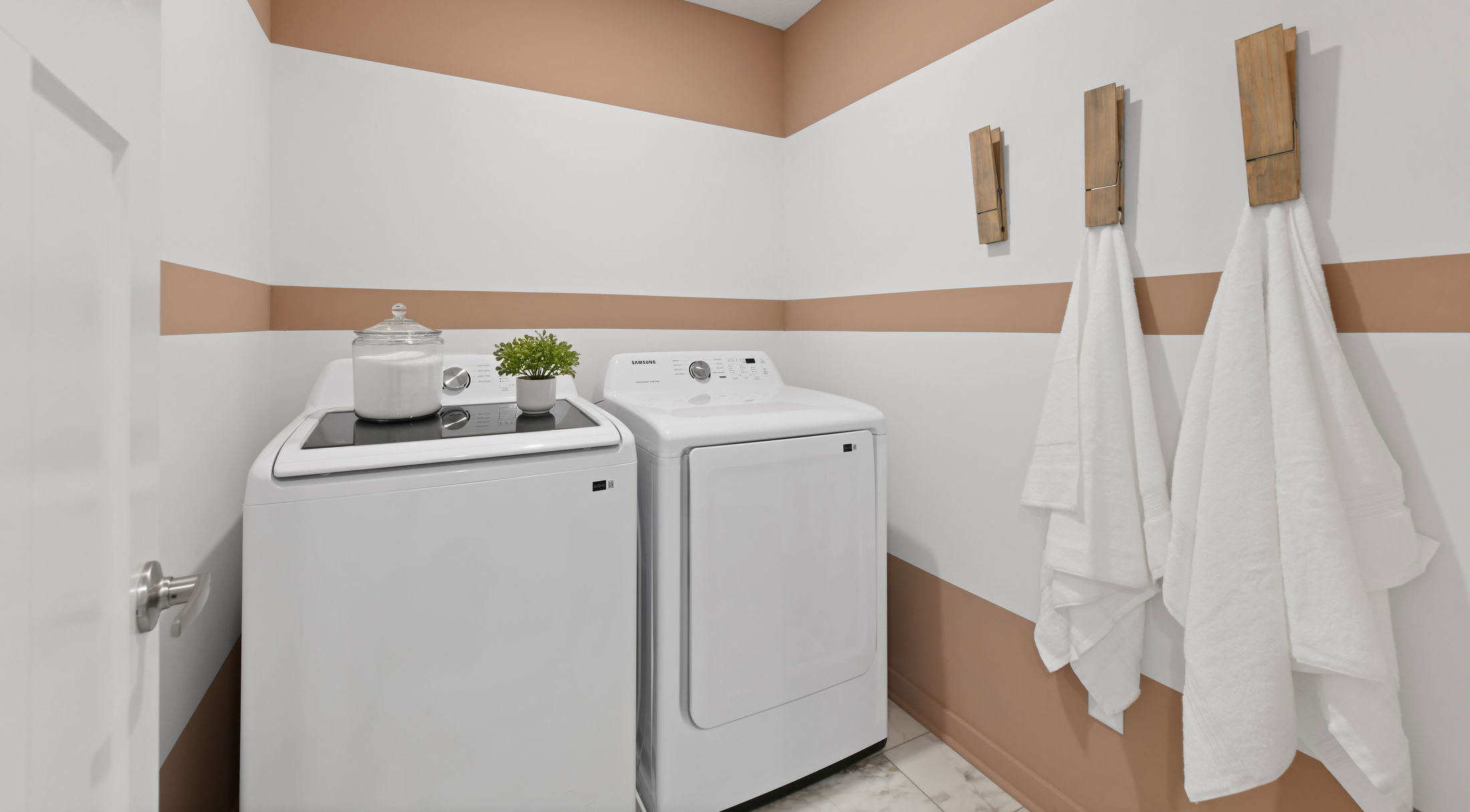 Modern laundry room with striped walls, featuring a washer and dryer set, topped with decorative items, and accented by wall hooks with white towels.
