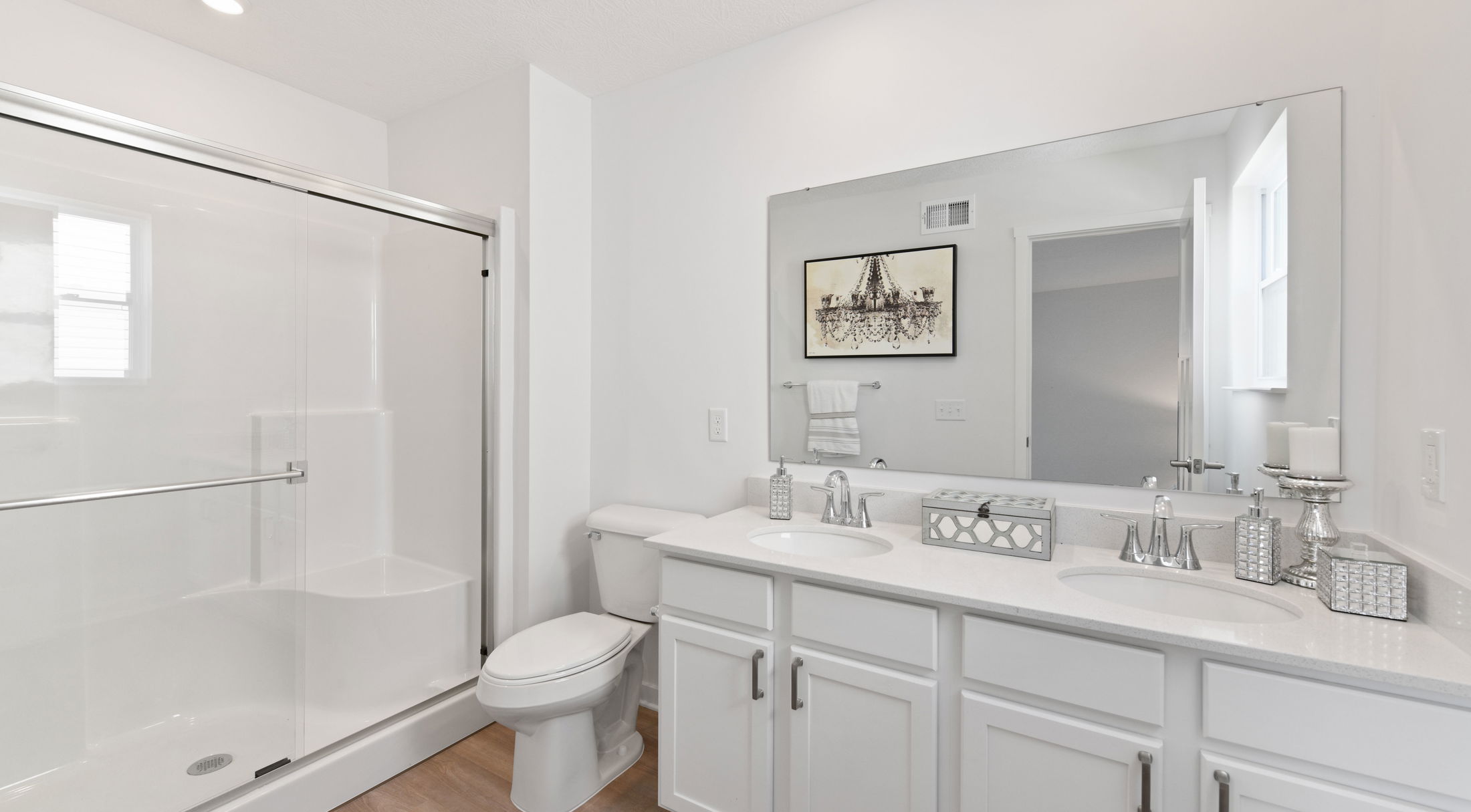 Clean and modern bathroom with a white double vanity, glass-enclosed shower, large mirror, and elegant wall art.