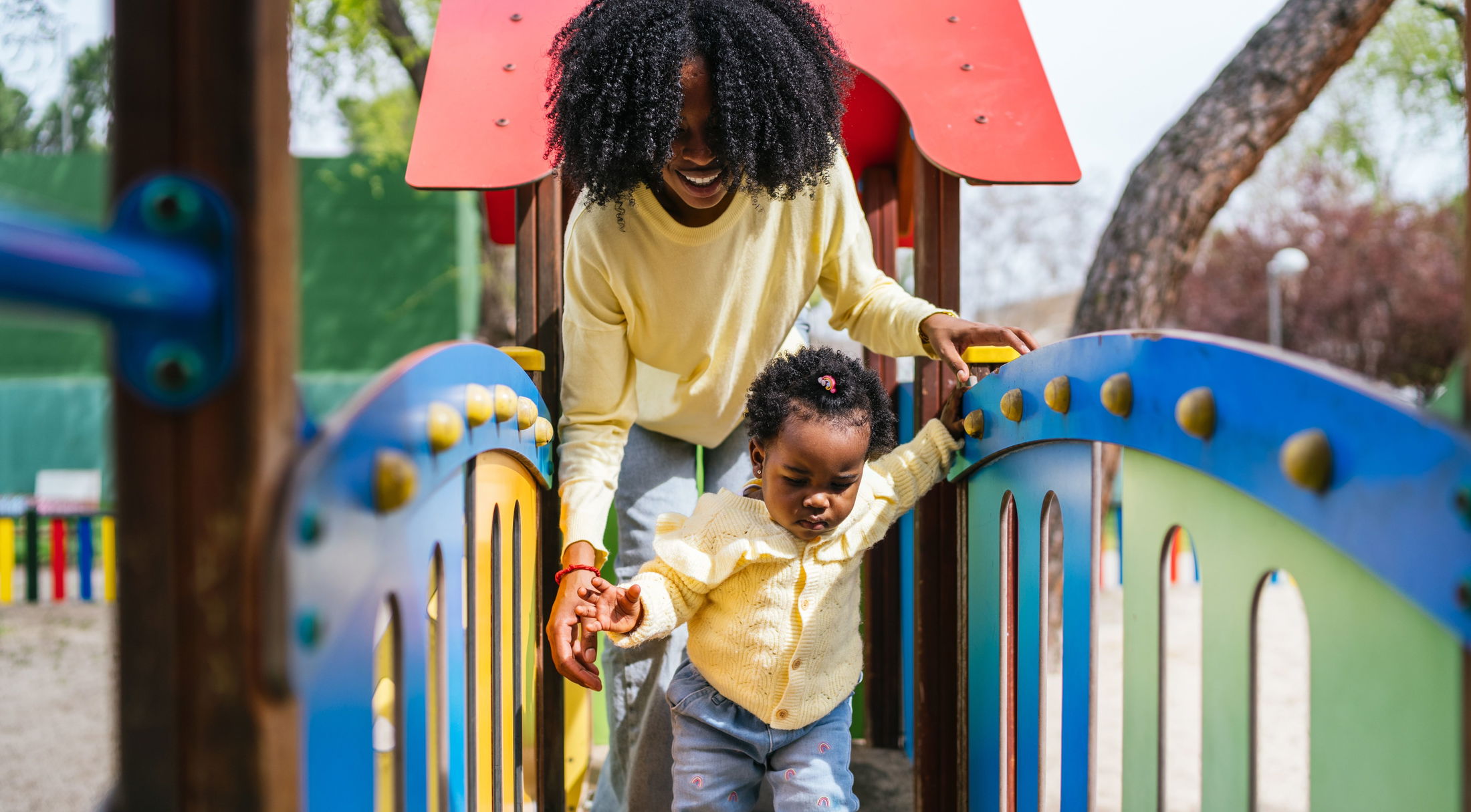 A smiling woman helps a young child walk across a colorful playground bridge under a sunny sky.