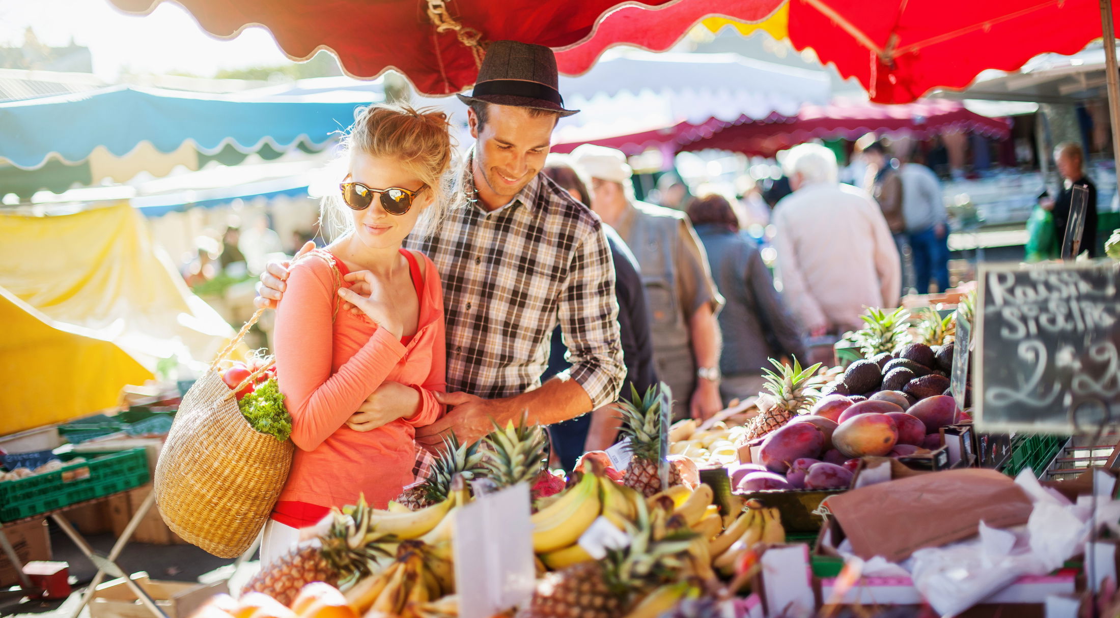 A couple enjoys selecting fresh fruits and vegetables at an outdoor market, under colorful canopies.