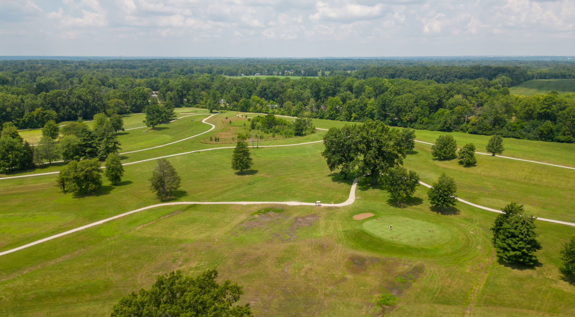 Aerial view of a lush, sprawling golf course surrounded by trees and winding pathways under a partly cloudy sky.