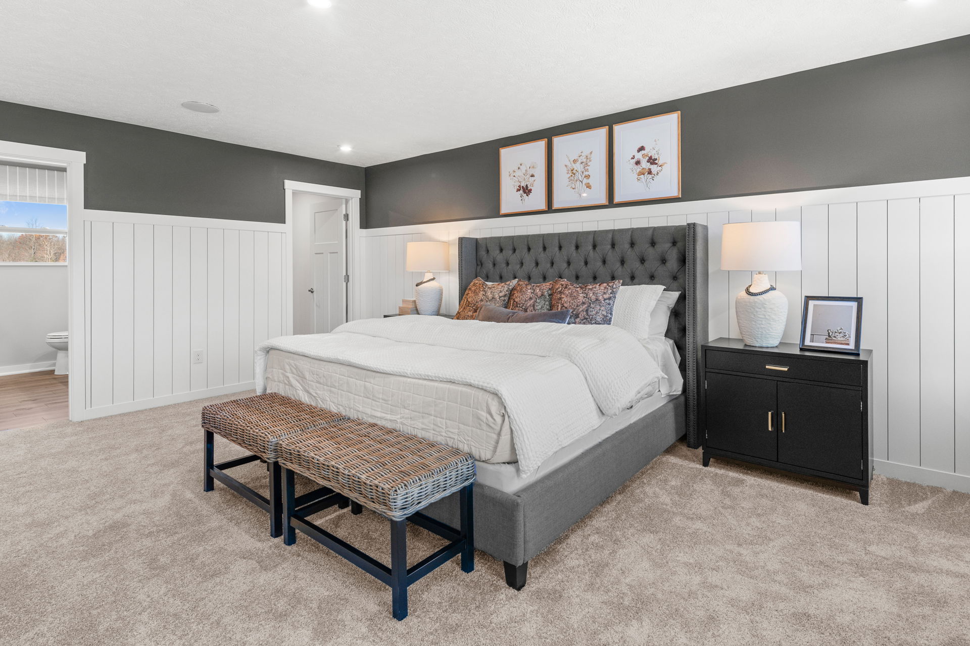 Elegant modern bedroom with plush gray upholstered bed, white bedding, woven benches, and stylish decor against gray and white paneled walls.