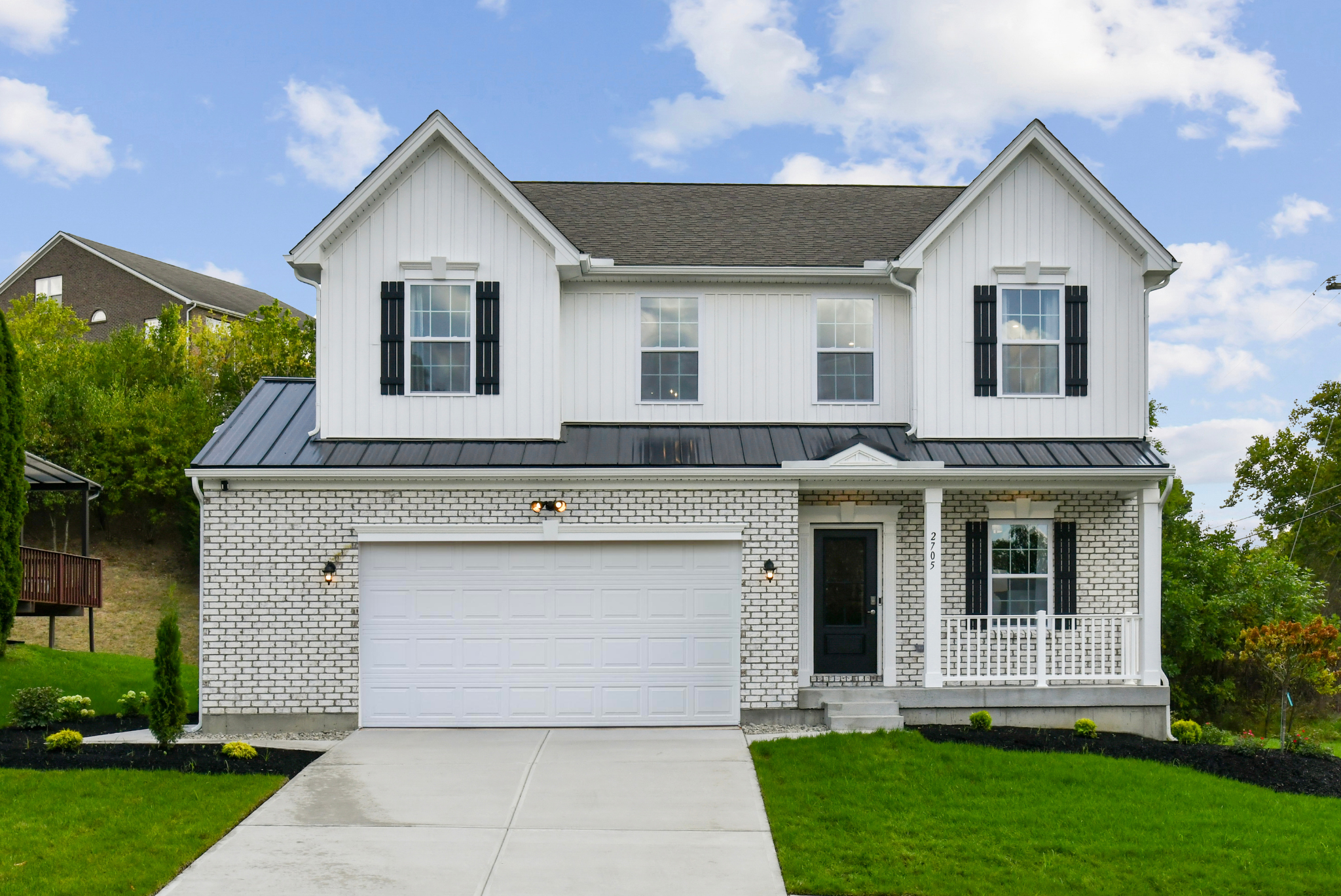 Front view of a modern two-story white house with black shutters, a metal roof, a garage, and a well-maintained lawn under a blue sky.