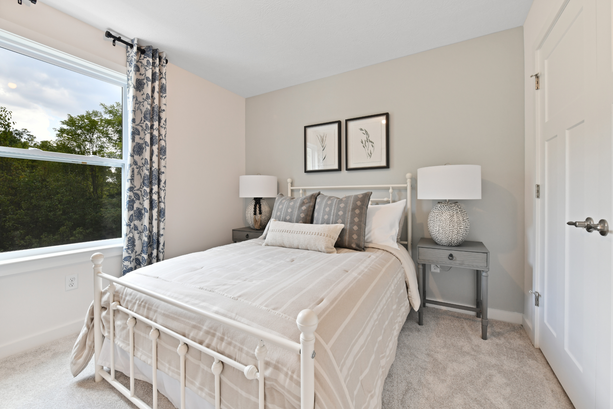 Cozy bedroom with beige and gray decor, featuring a neatly made bed, patterned curtains, and framed botanical artwork by a window overlooking greenery.