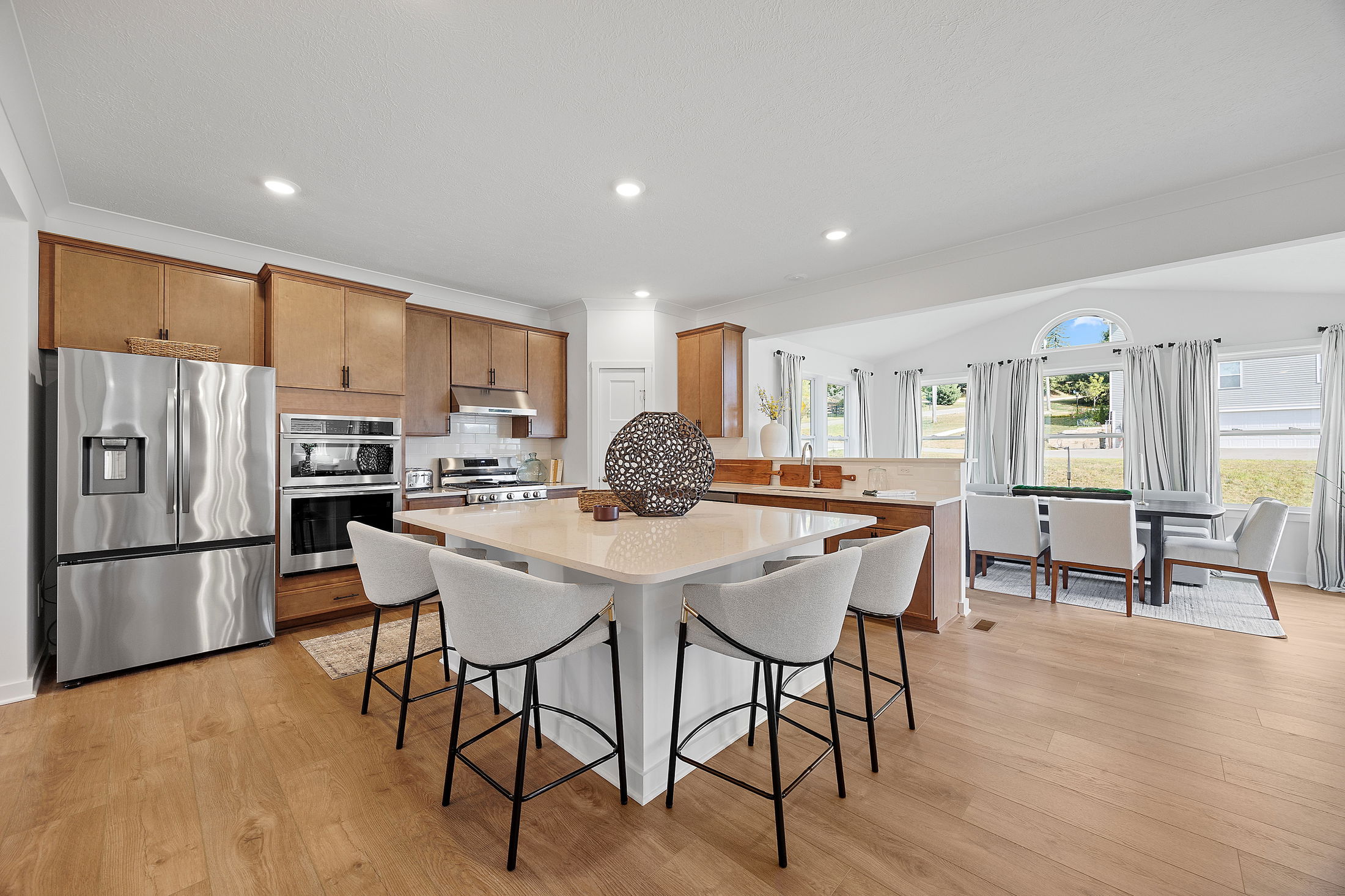 Modern kitchen with wooden cabinetry, stainless steel appliances, a central island with bar stools, and a dining area in an open, sunlit space.
