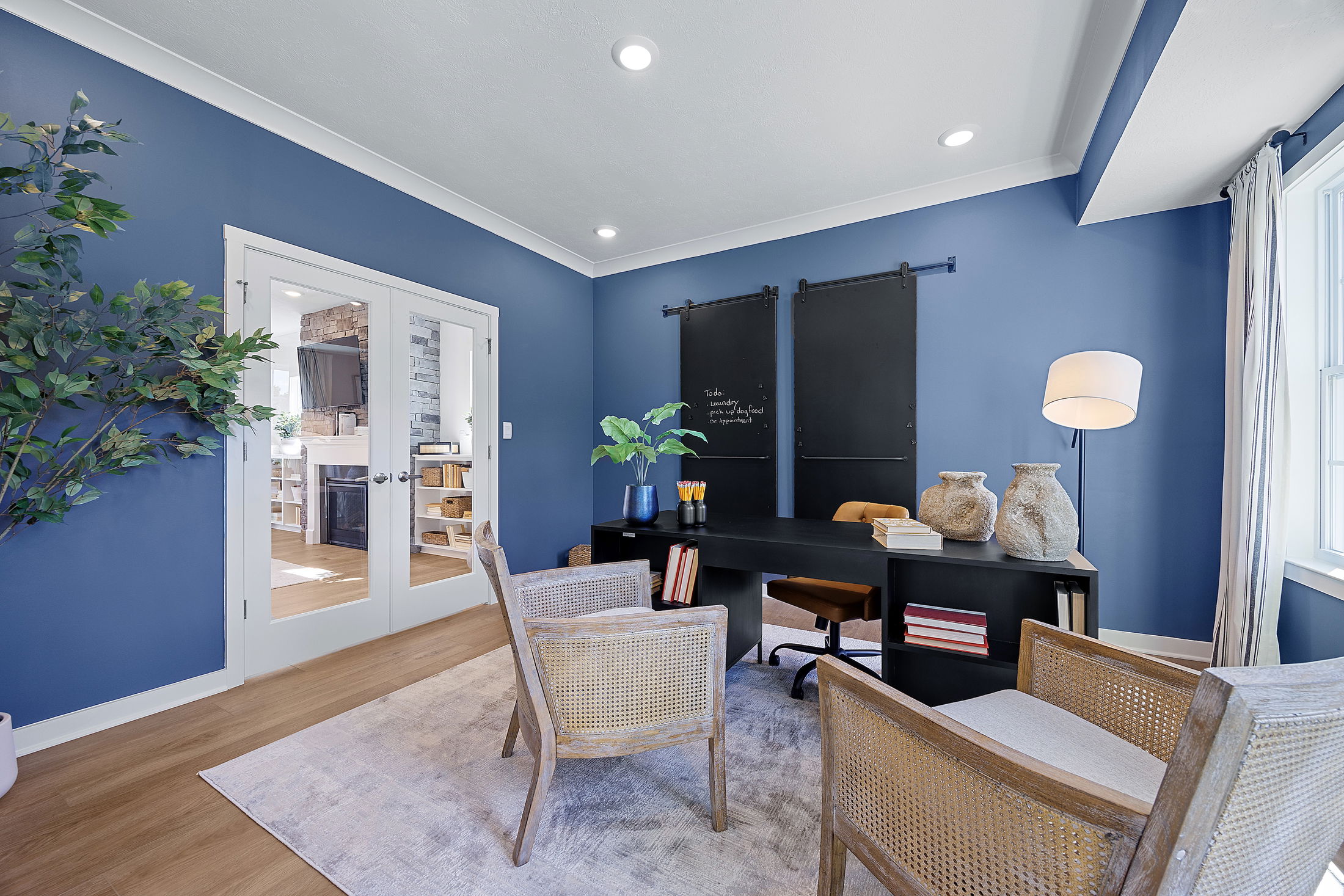 Modern home office with blue walls, wooden furniture, large windows, and decorative vases on a sleek black desk.