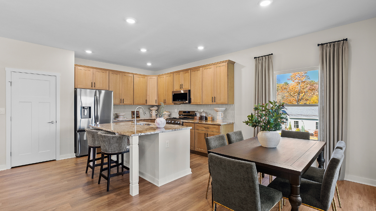 Modern kitchen with wooden cabinets, center island with granite countertop, stainless steel appliances, dining table, and large window with a scenic view.